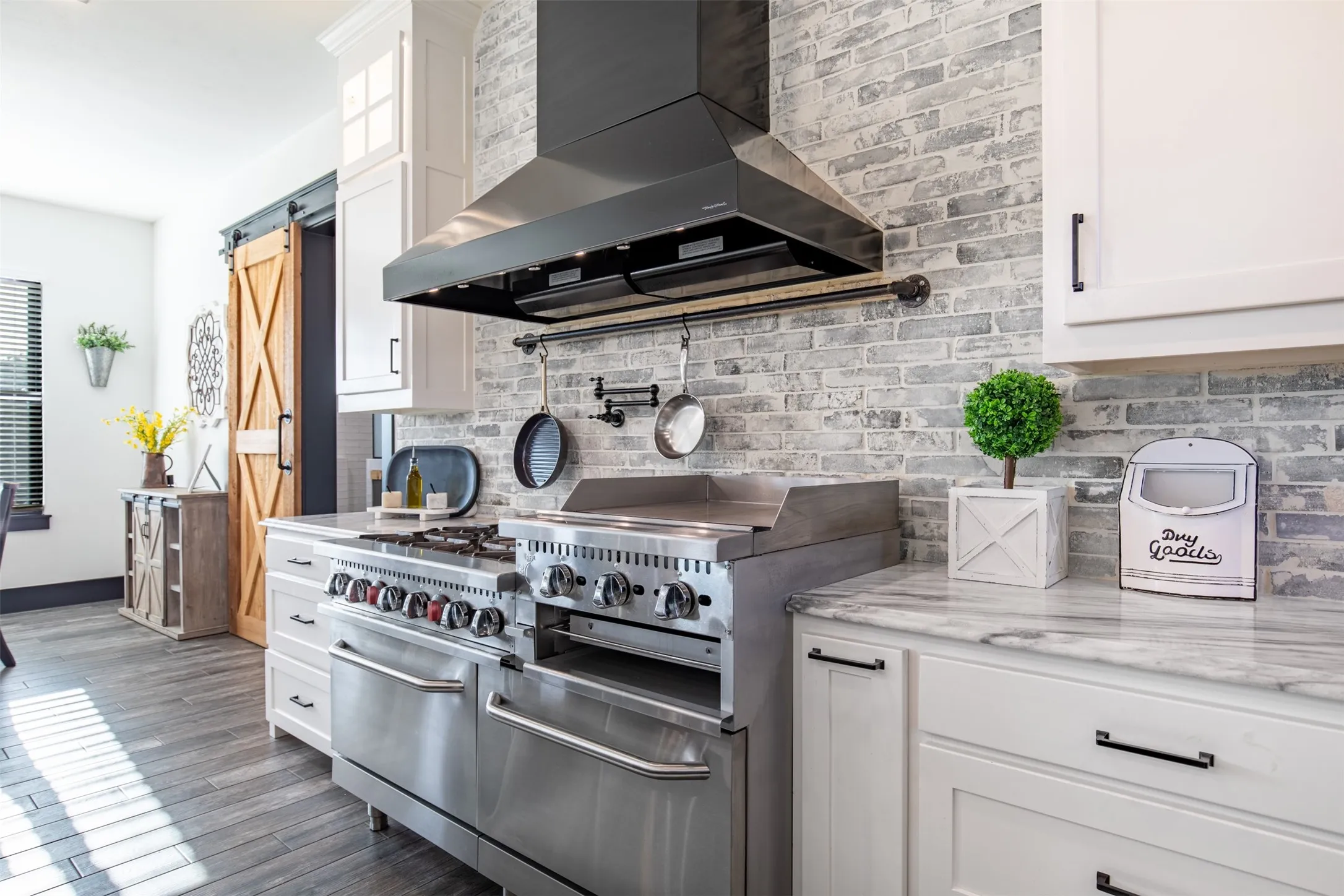 Professional-grade stainless steel range with a griddle, black range hood, light gray brick brick backsplash, white cabinetry with black hardware, and wood-finish flooring