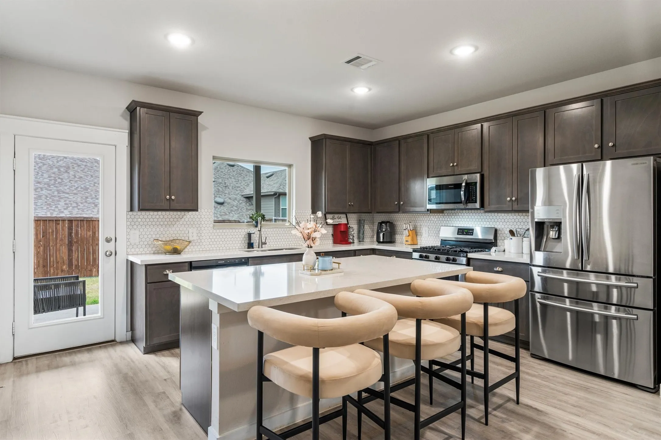 Modern kitchen featuring dark wood-finish cabinetry, stainless steel appliances, a white hexagon tile backsplash, and a large central island with a light-toned countertop