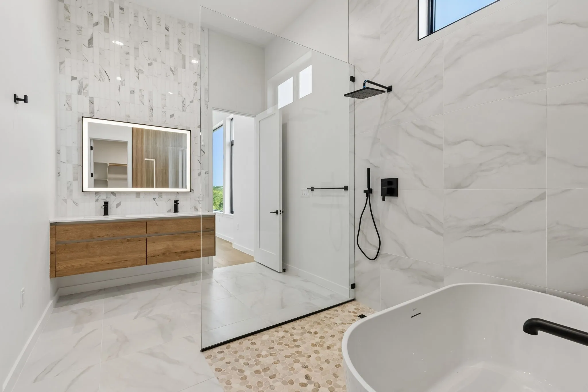 Underlit floating dual vanity with integrated sinks, illuminated mirror, glassed wet room with shower, freestanding soaking tub, and floor-to-ceiling tile