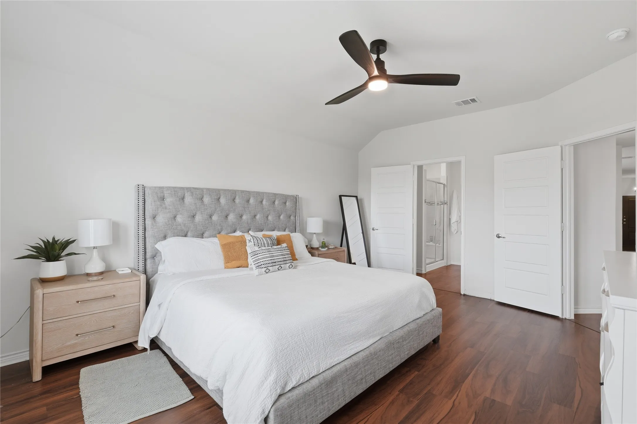 Spacious room featuring rich wood-finish flooring, a light gray tufted headboard, and a contemporary ceiling fan