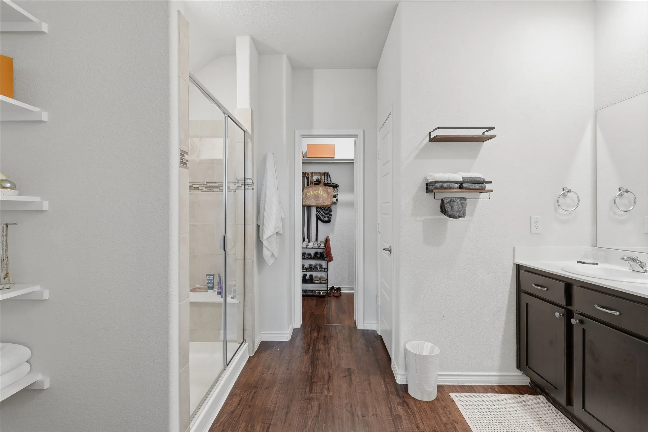 Spacious bathroom featuring wood-finish flooring, a glass-enclosed shower, dark wood vanity with a white countertop, and built-in shelving