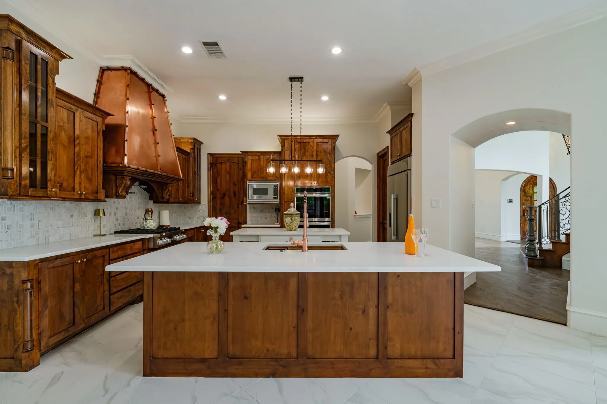 Gourmet kitchen featuring a large central island with white countertop, rich wood cabinetry, stainless steel appliances, a distinctive copper range hood, and white tile flooring