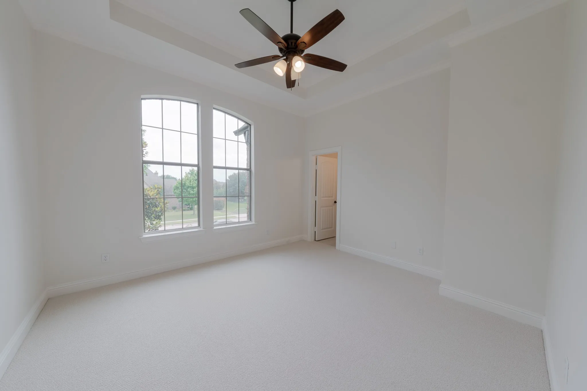 Spacious room with light neutral carpet flooring, arched windows, a tray ceiling, and a ceiling fan with light fixture