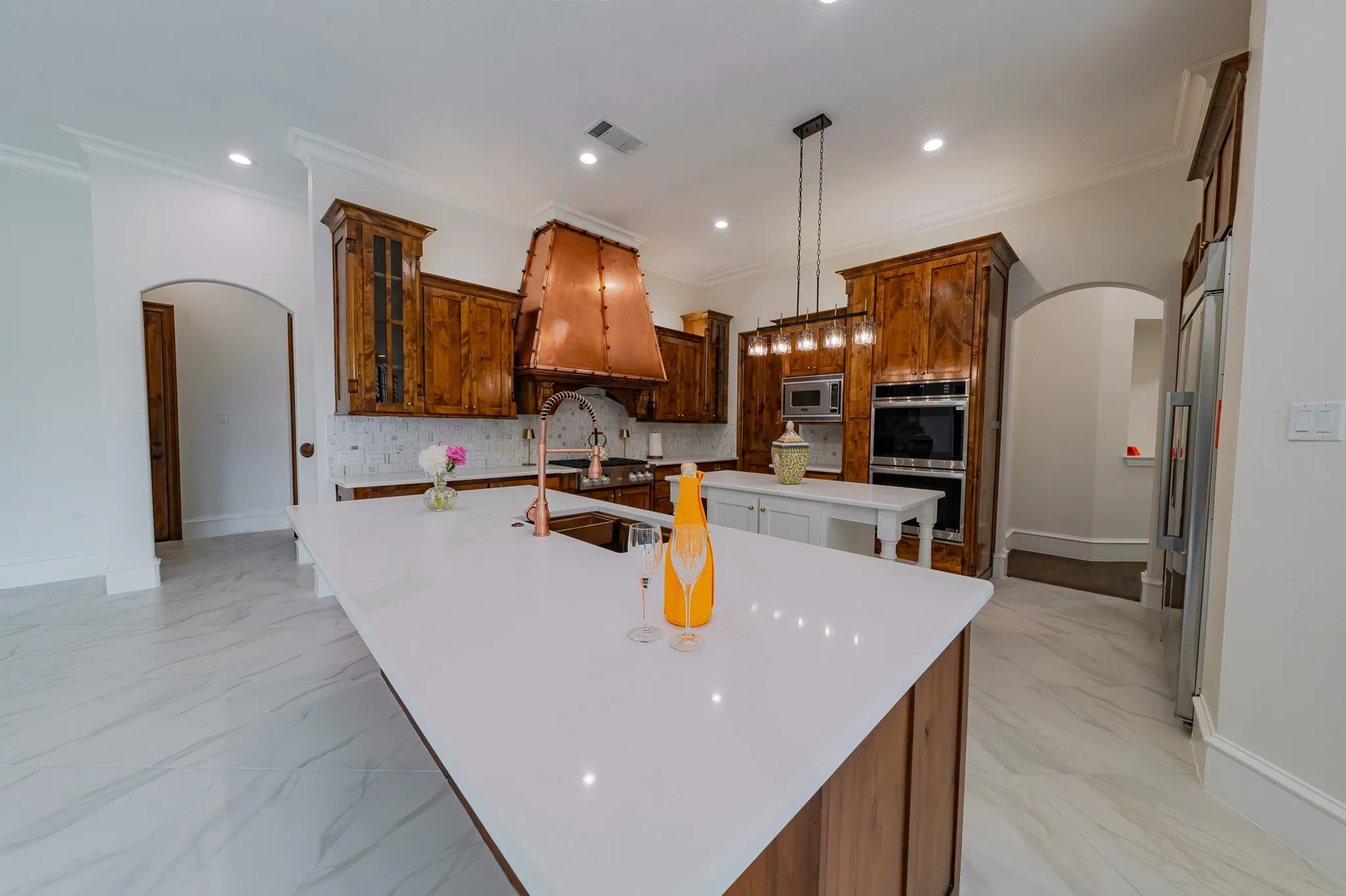 Gourmet kitchen featuring a large central island with a white quartz countertop, rich wood-finish cabinetry, a copper range hood, stainless steel double ovens, and large format marble-look tile flooring