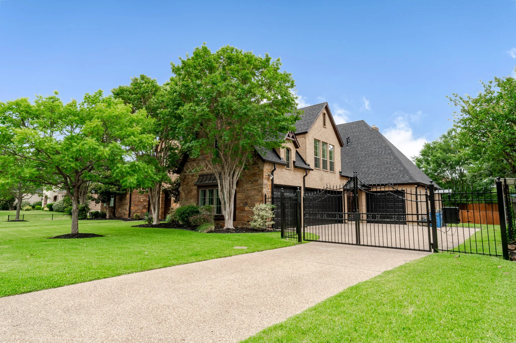 Gated driveway entry leading to a multi-4car garage, complemented by a stone and stucco exterior, dark shingle roof, and mature landscaping