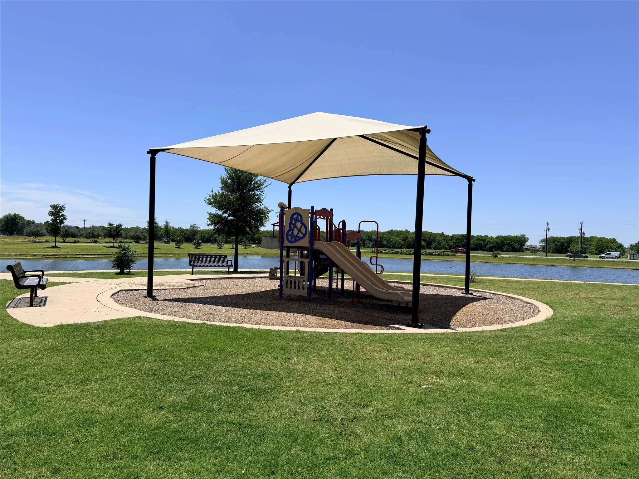 Playground structure with shade canopy, adjacent to a pond with a concrete pathway and green space