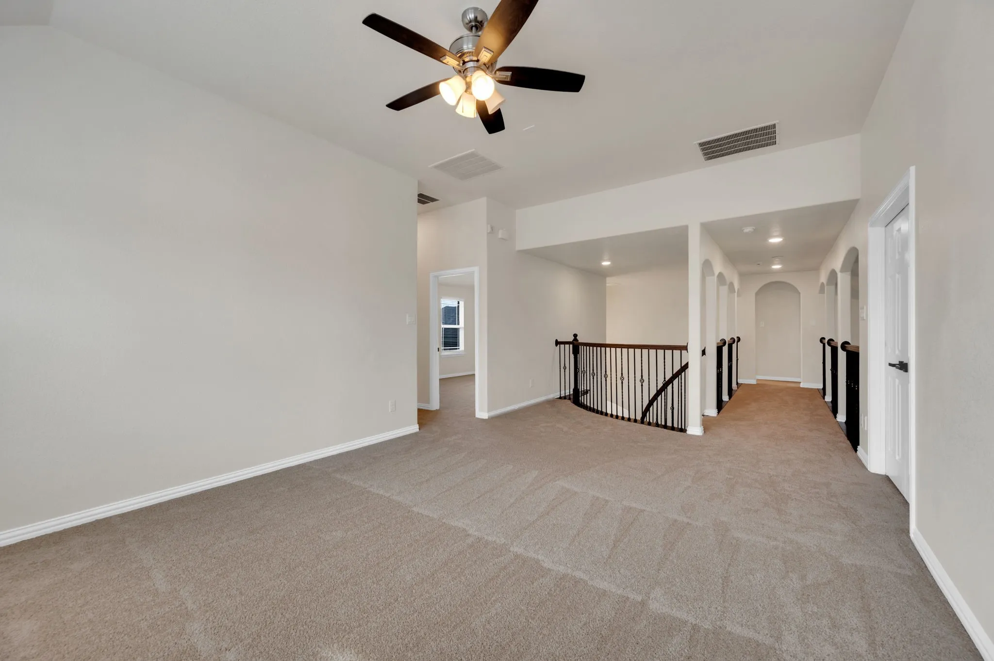 Carpeted open area featuring a ceiling fan with integrated lighting, a curved black metal railing overlooking a lower level, and an arched hallway with recessed lighting