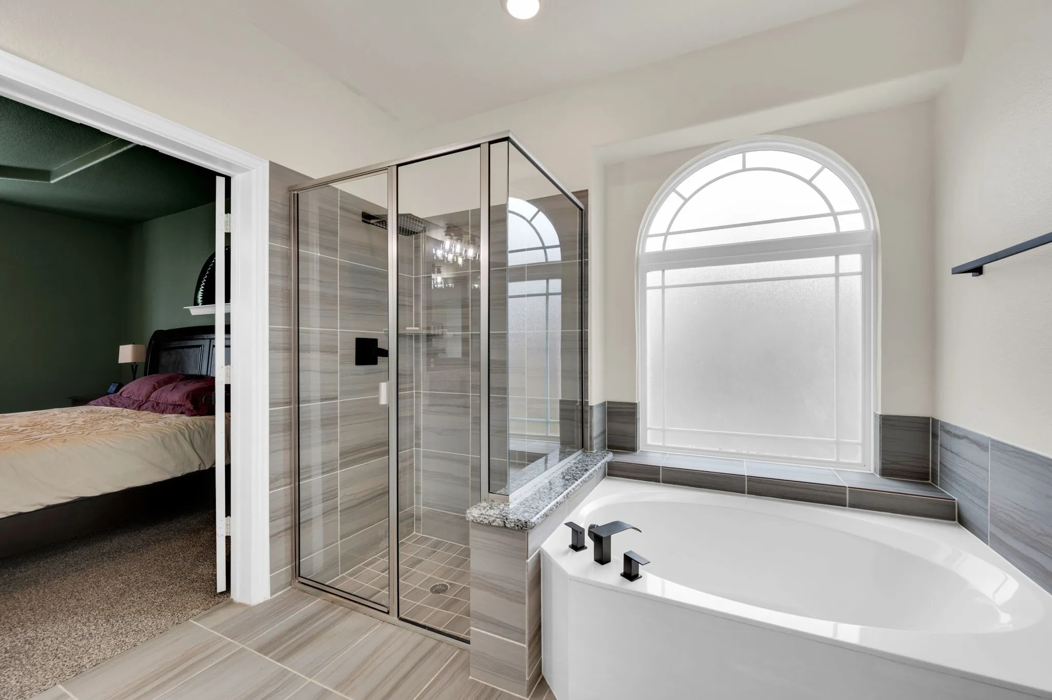 Bathroom featuring a tiled corner soaking tub, a frameless glass shower enclosure, a large arched window with privacy glass, and tile flooring