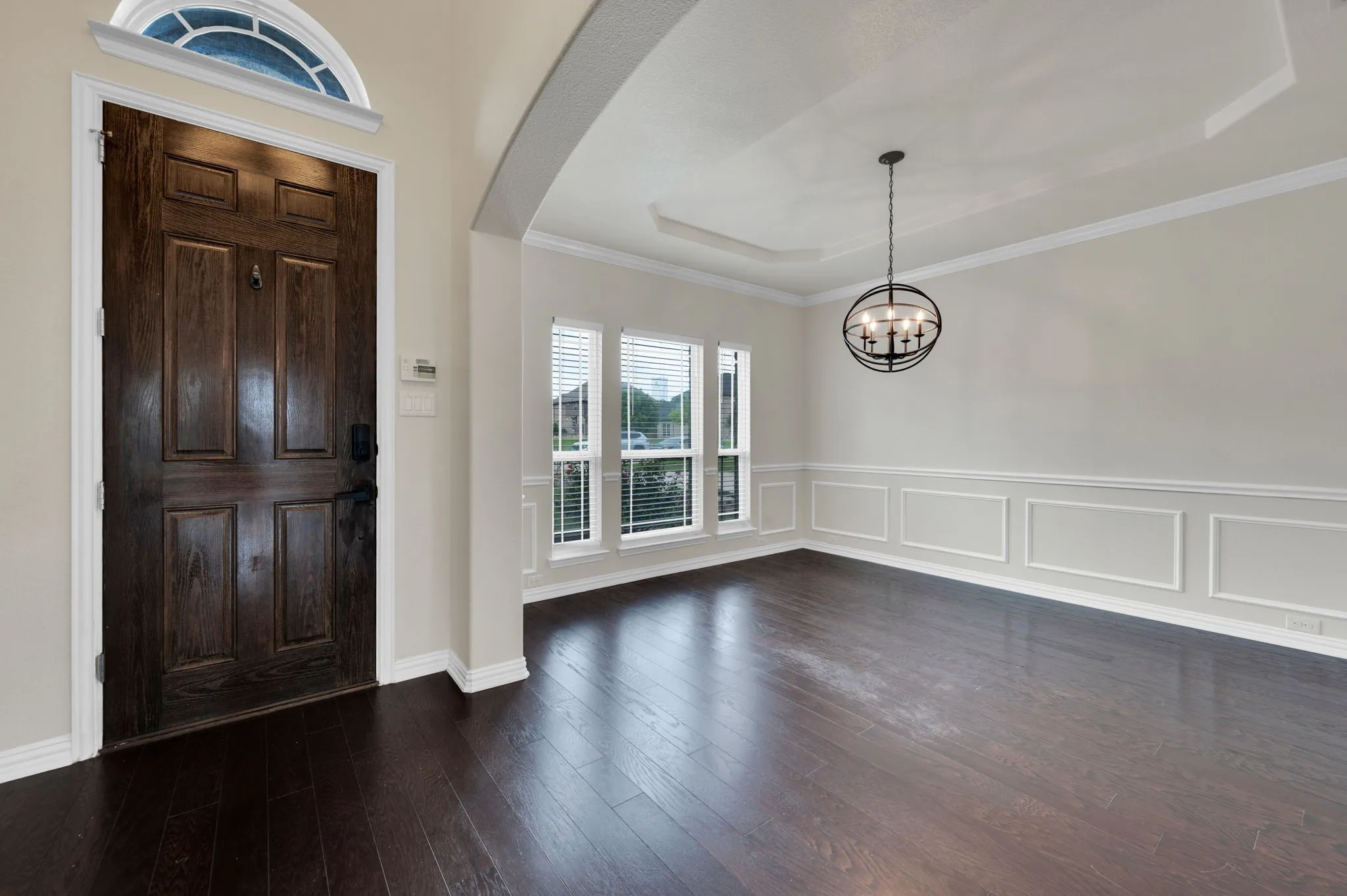 Rich wood-finish flooring complements the detailed paneling and tray ceiling in this inviting space, illuminated by an elegant orb chandelier and natural light from multiple windows