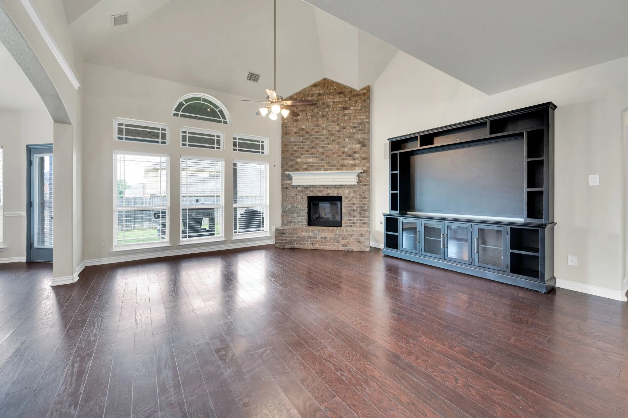 Spacious living area featuring high vaulted ceilings, expansive wood-finish flooring, and a brick fireplace with a white mantel