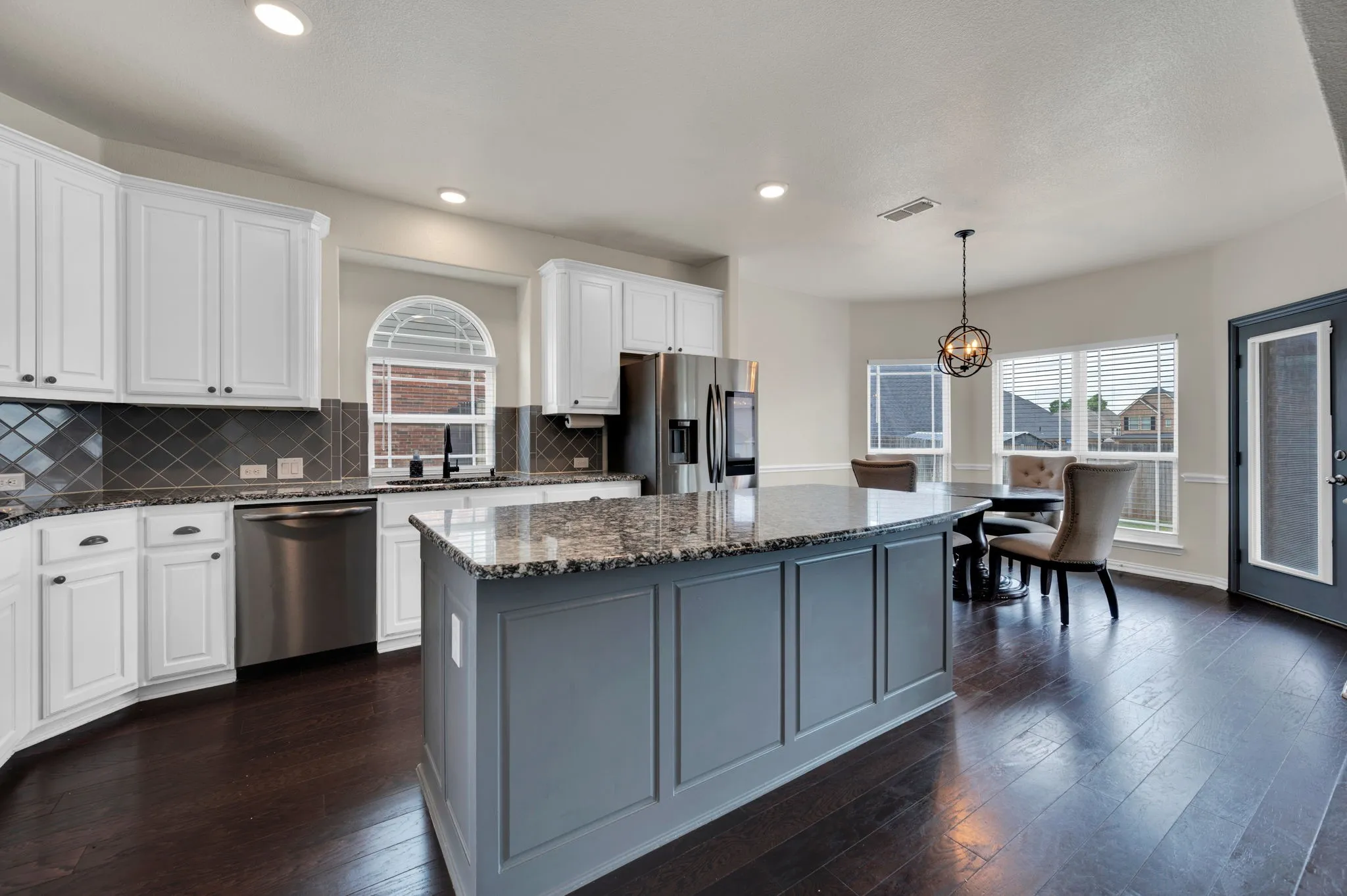 Spacious kitchen featuring an expansive center island with stone countertop, white cabinetry, stainless steel appliances, dark wood-finish flooring, and a dining nook