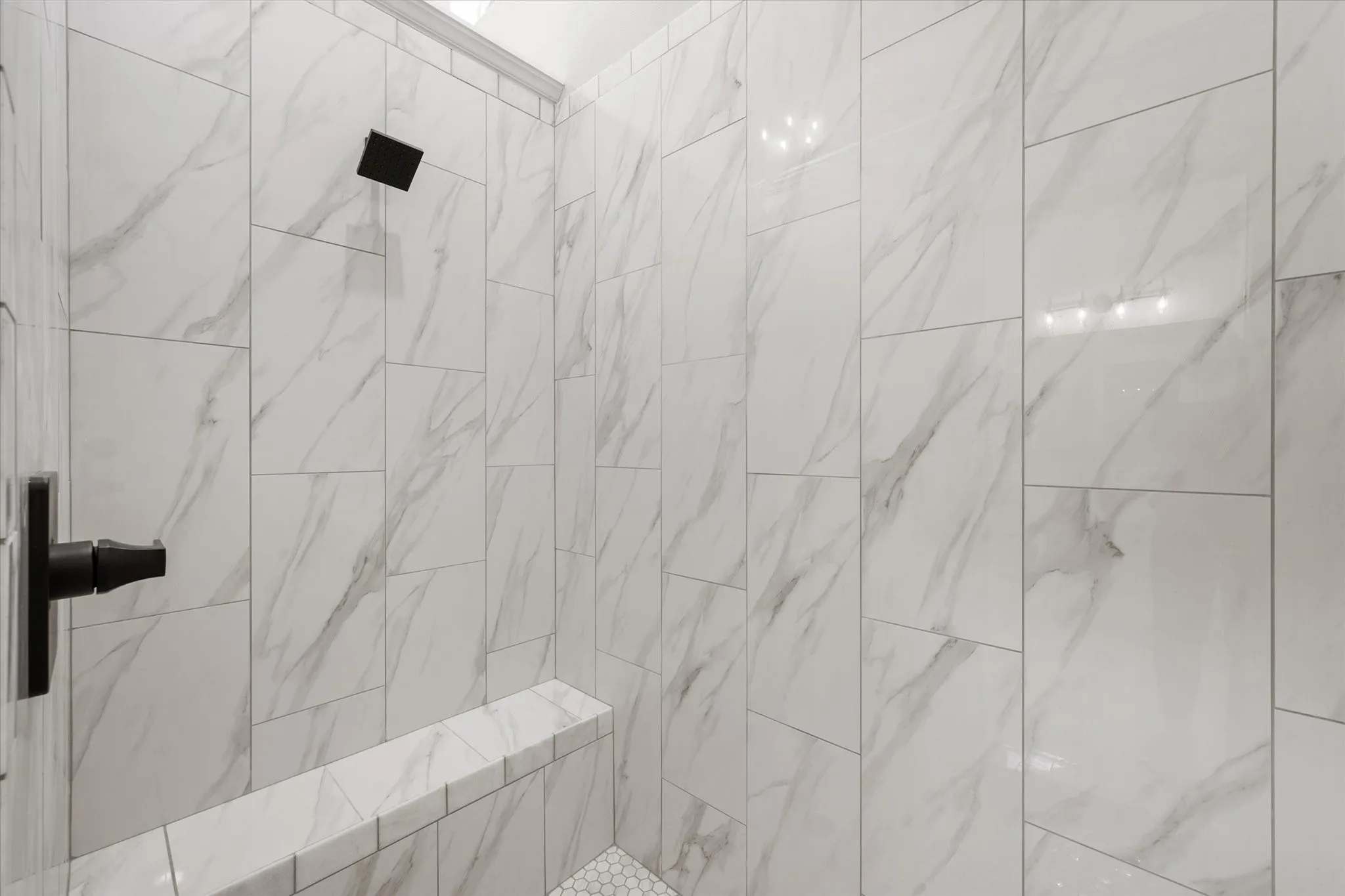 Shower enclosure featuring floor-to-ceiling white marble-look tile, a built-in bench, matte black showerhead, and matte black lever handle