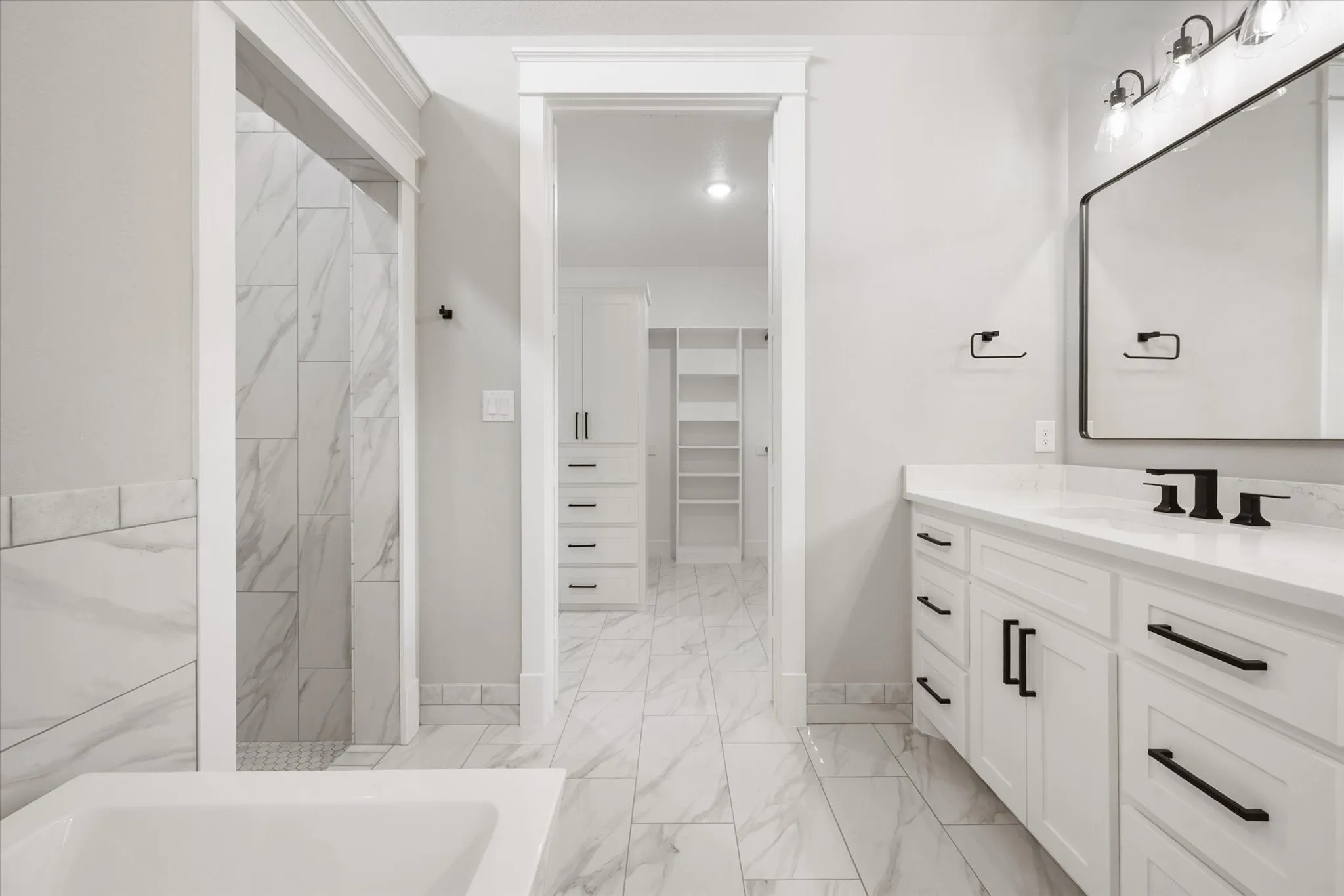 Spacious bathroom featuring a double vanity with white cabinetry and black hardware, a large rectangular mirror with black frame, and a tub with marble-look tile surround