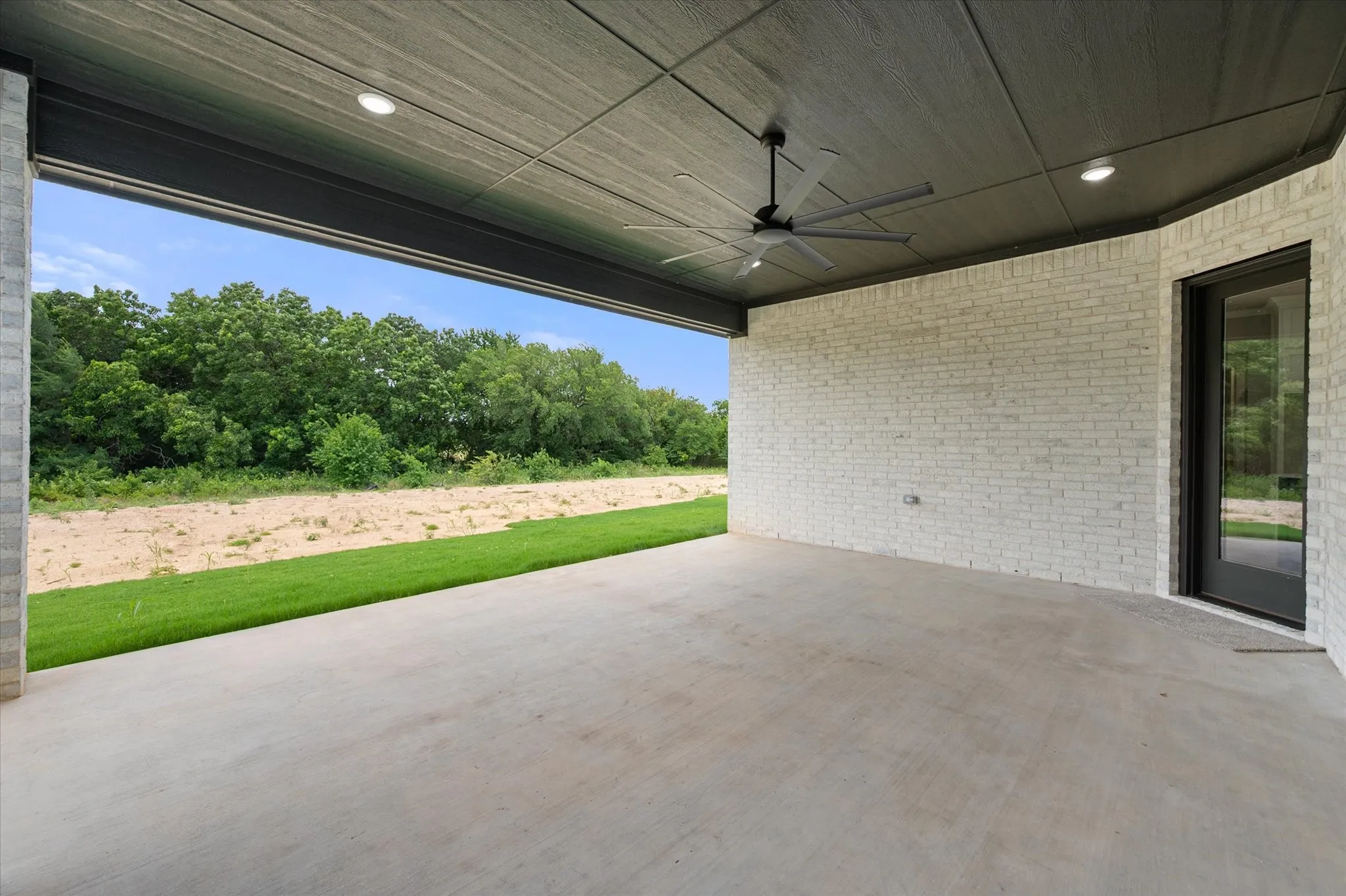 Expansive covered patio featuring a concrete surface, white brick wall, and a dark wood-finish ceiling with recessed lighting
