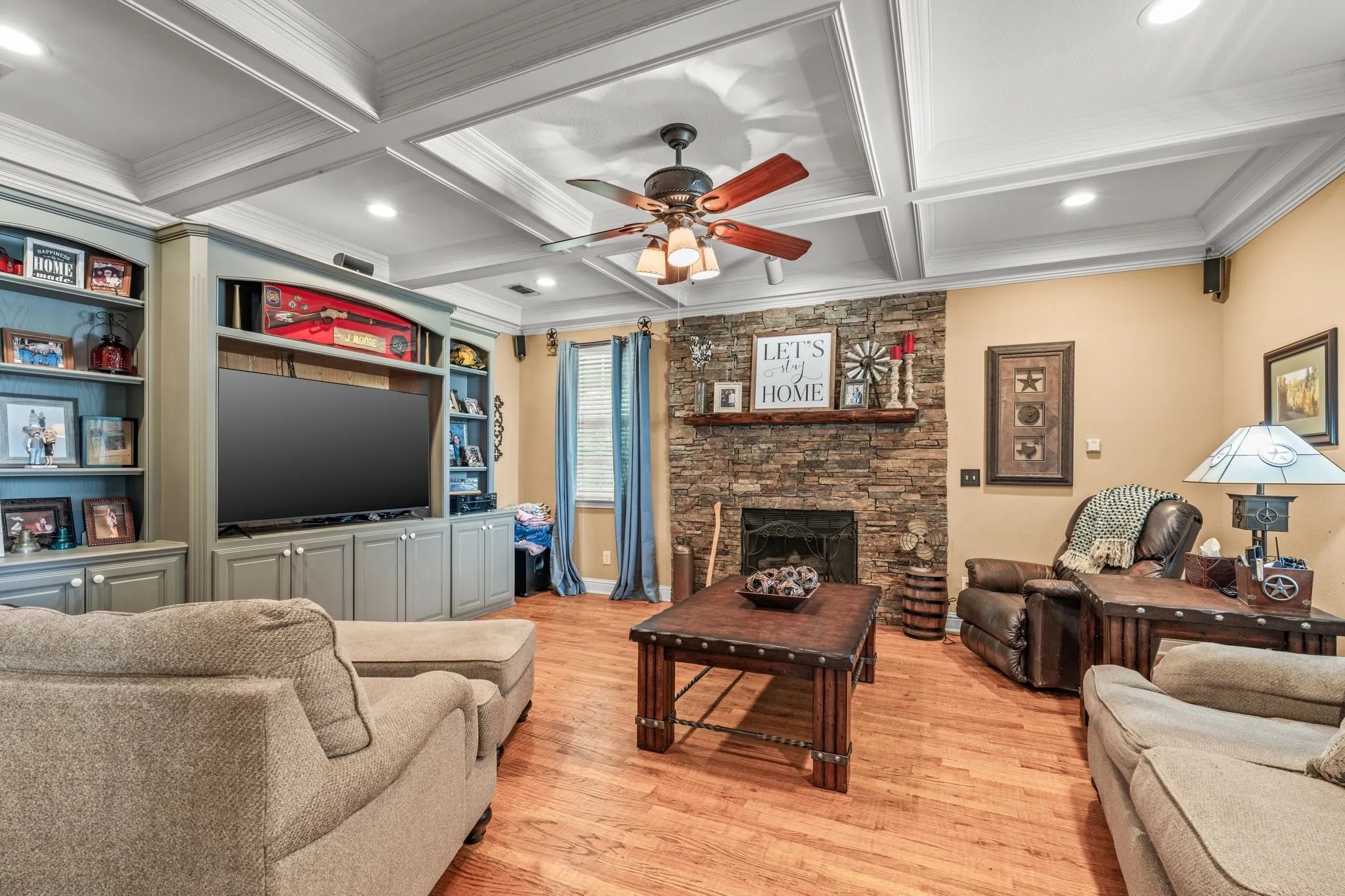Living area featuring rich wood-finish flooring, a stone-clad fireplace with a wood mantel, and a detailed coffered ceiling with a ceiling fan