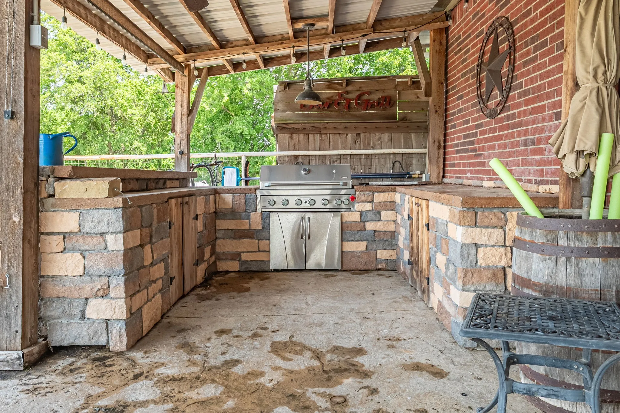 Covered outdoor kitchen featuring a built-in stainless steel grill, stone veneer cabinetry, wood support beams, and a corrugated metal roof