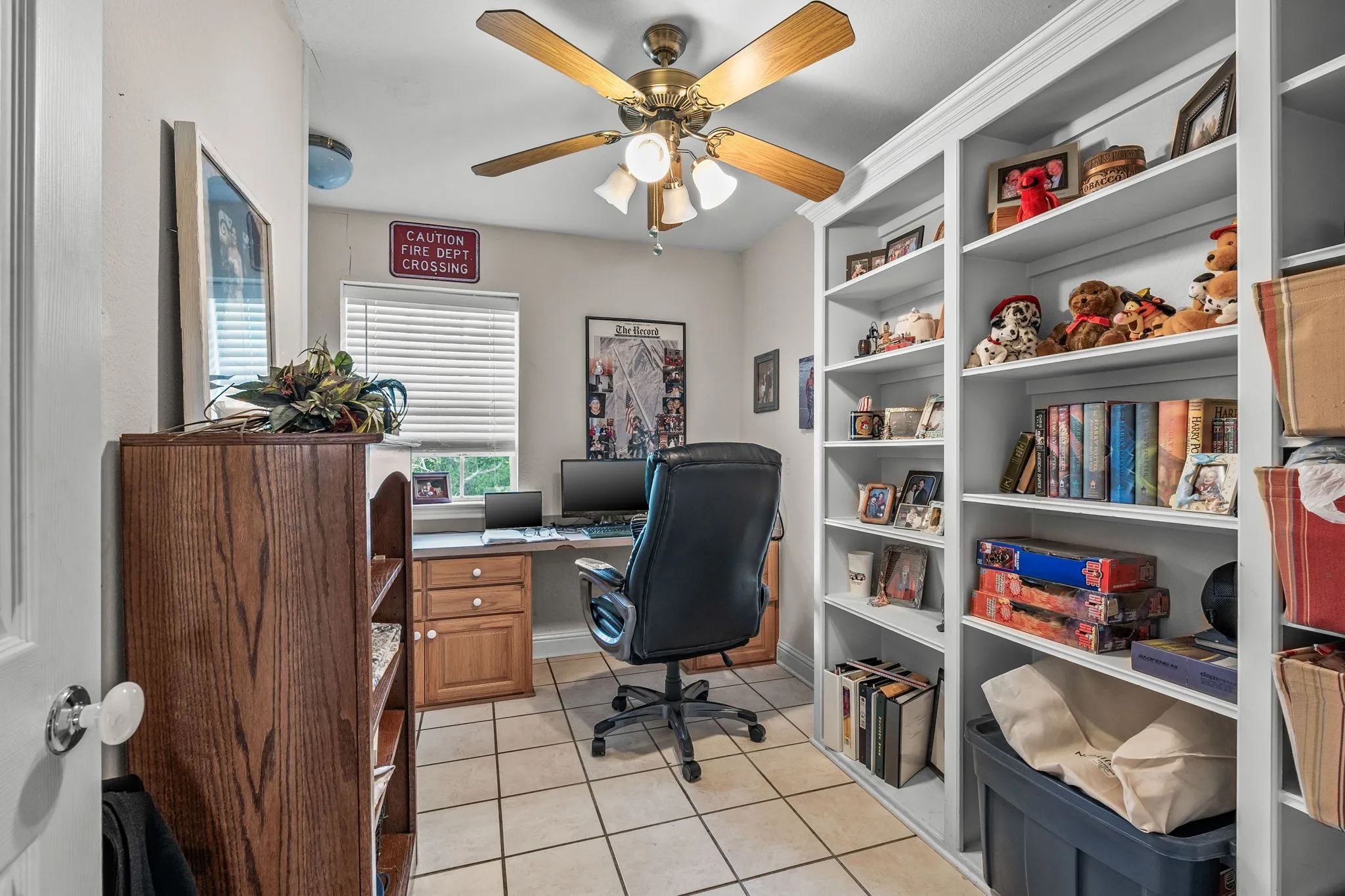 Dedicated room featuring built-in white shelving, wood-finish desk cabinetry, a window with horizontal blinds, a ceiling fan with light fixture, and tiled flooring