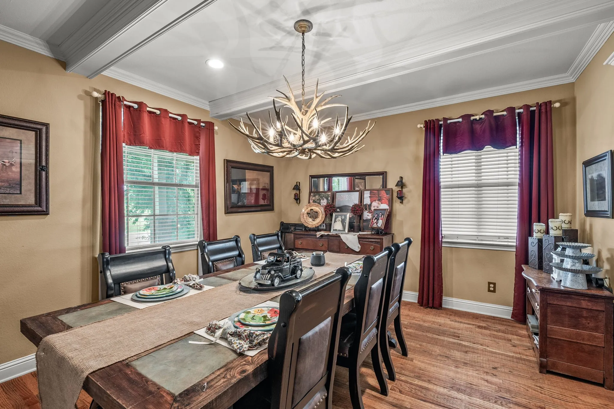 Dining area featuring a decorative antler chandelier, wood-finish flooring, and two windows with blinds and red curtains