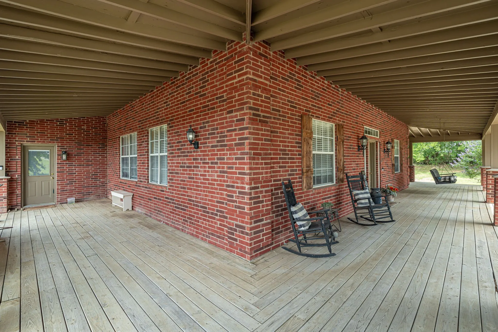 Expansive covered porch featuring a red brick exterior, wood-finish flooring, multiple windows with white blinds, and decorative wall sconces