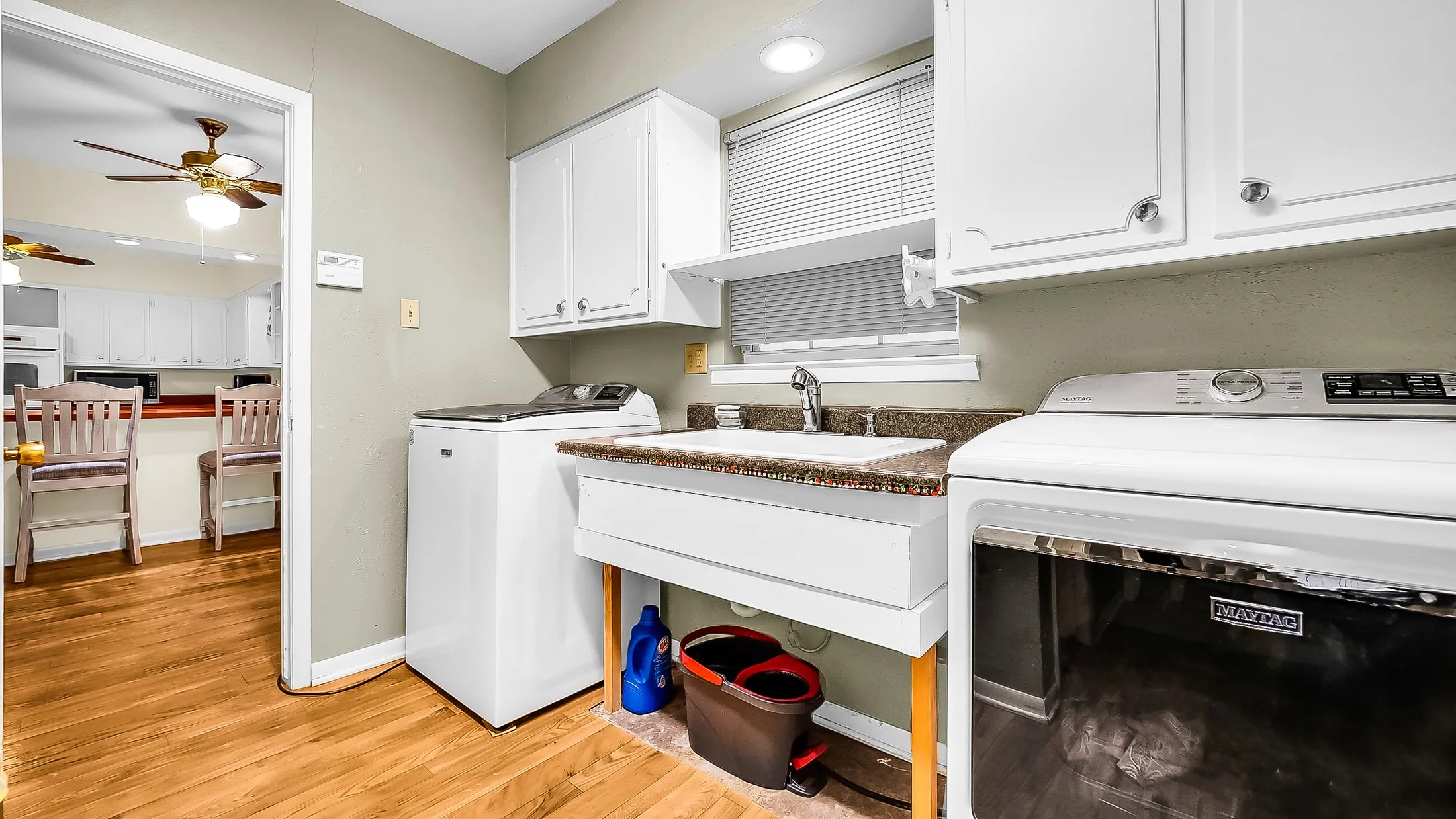 Dedicated laundry area featuring wood-finish flooring, white cabinetry, a utility sink, and a window with blinds