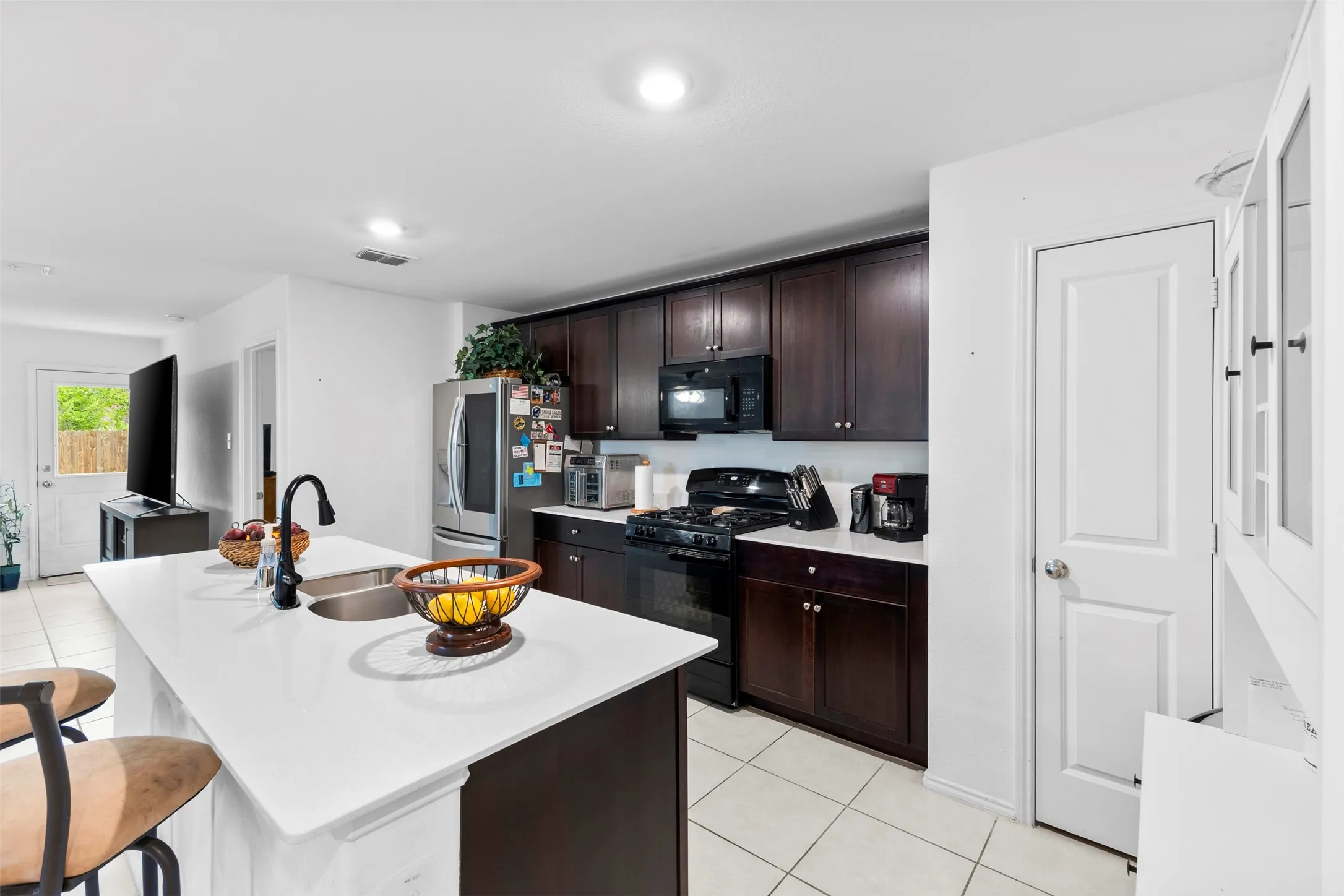 Spacious kitchen featuring an island with a double basin sink and a black gooseneck faucet, dark wood-finish cabinetry, light countertops, stainless steel refrigerator, and tile flooring