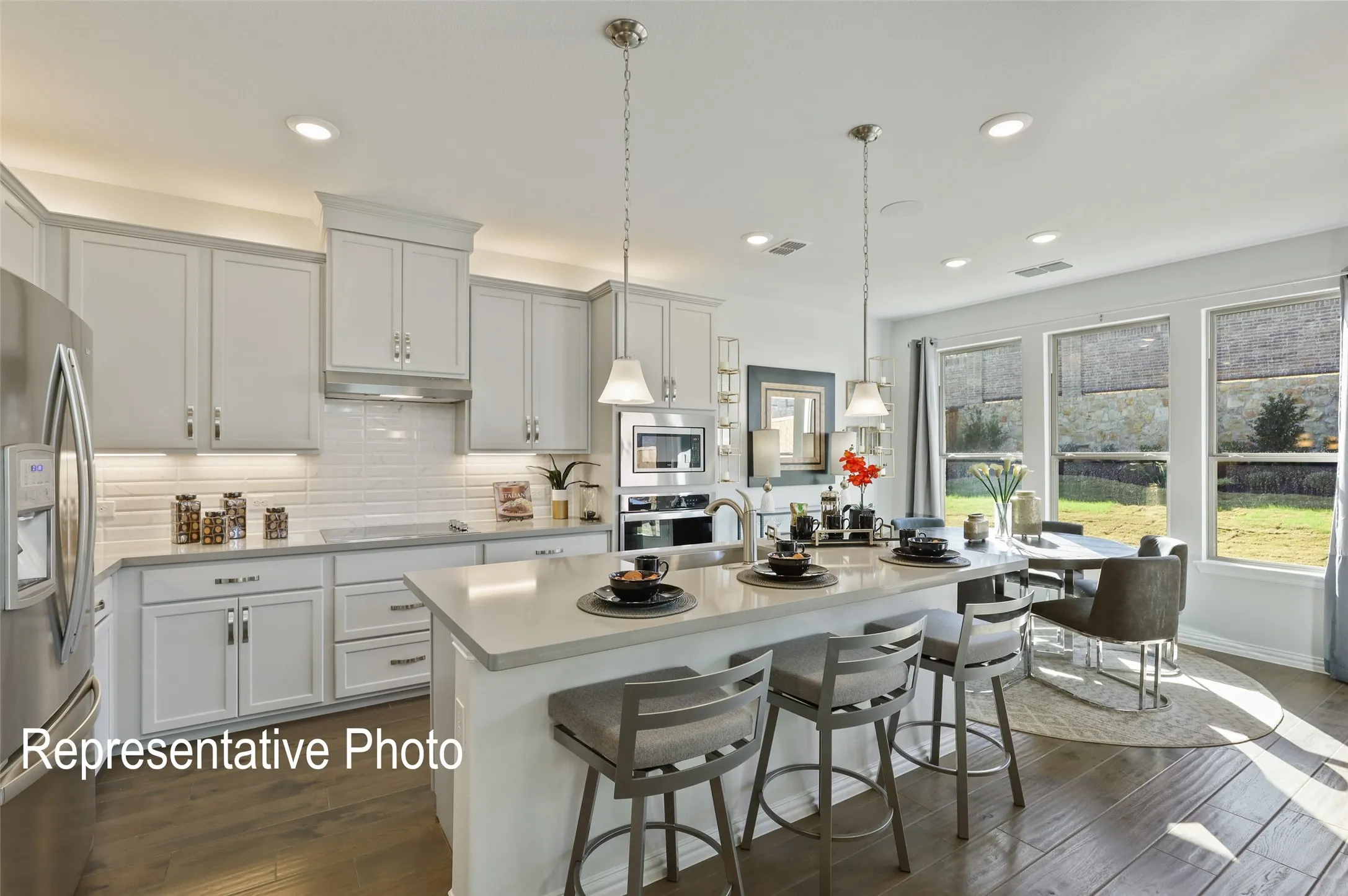 Open-concept kitchen featuring light-toned cabinetry, stainless steel appliances, a center island with pendant lighting, wood-finish flooring, and a dining area with multiple windows