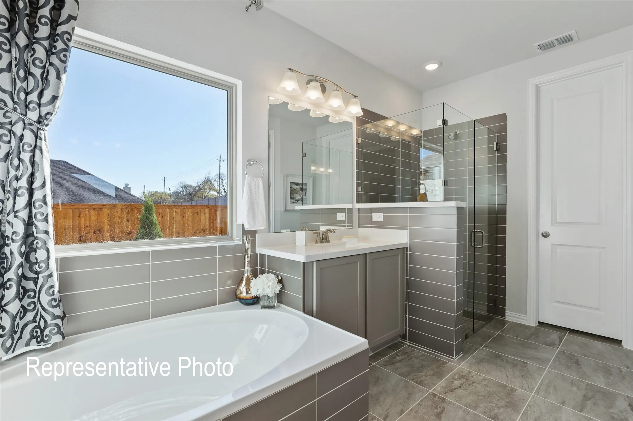 Spacious bathroom featuring a soaking tub, separate glass-enclosed shower, a vanity with a countertop, gray tiled walls, and large format floor tiles