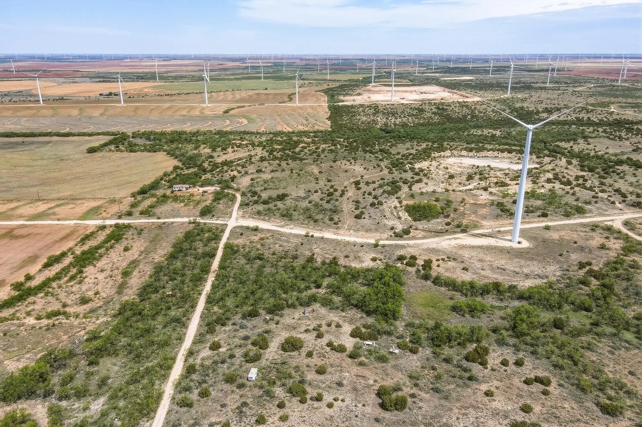 Expansive rural landscape featuring a network of dirt roads, scattered green vegetation, and numerous wind turbines across the horizon