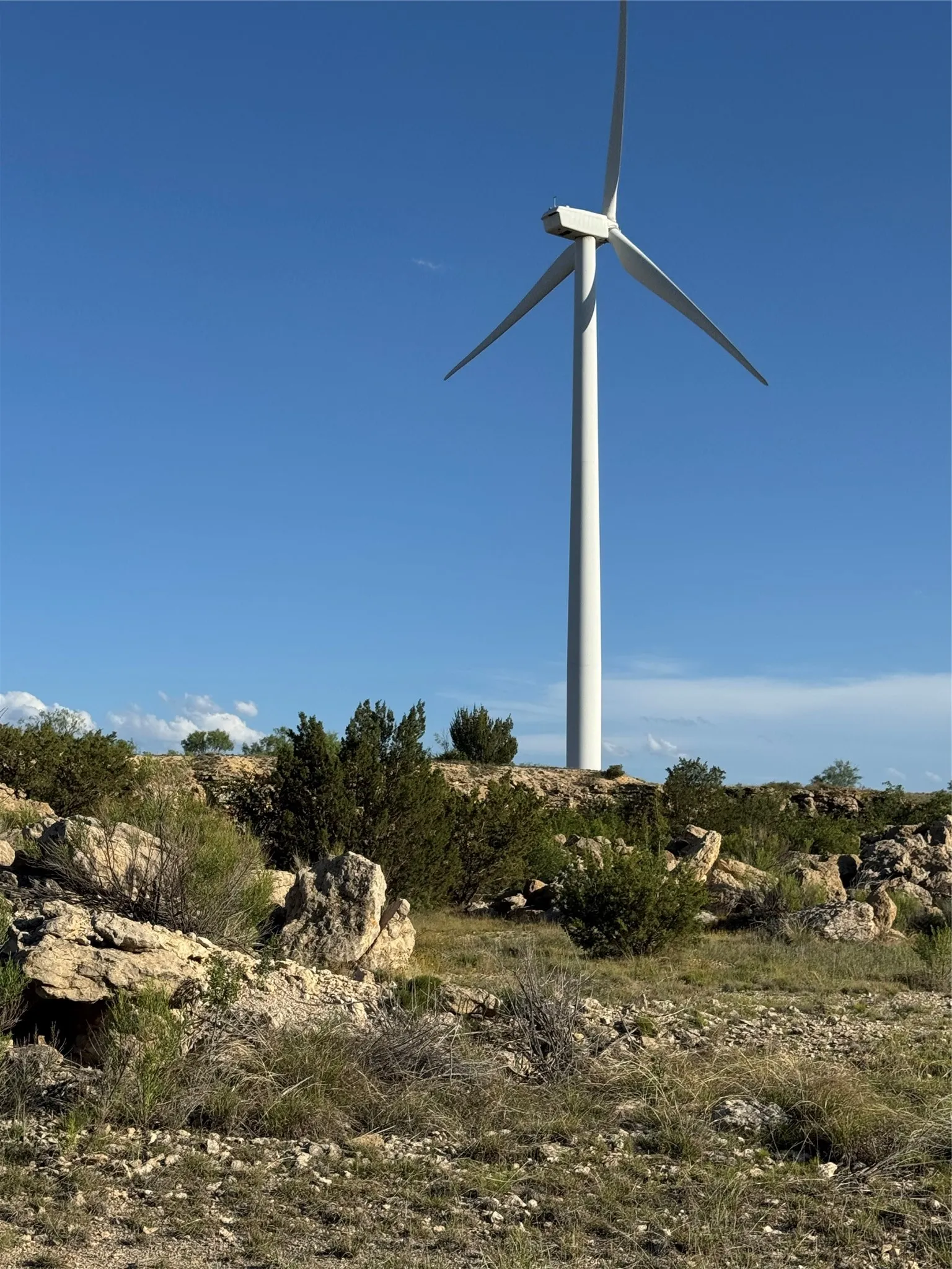 Towering wind turbine set against a clear blue sky, surrounded by natural rock formations, sparse vegetation, and rugged terrain