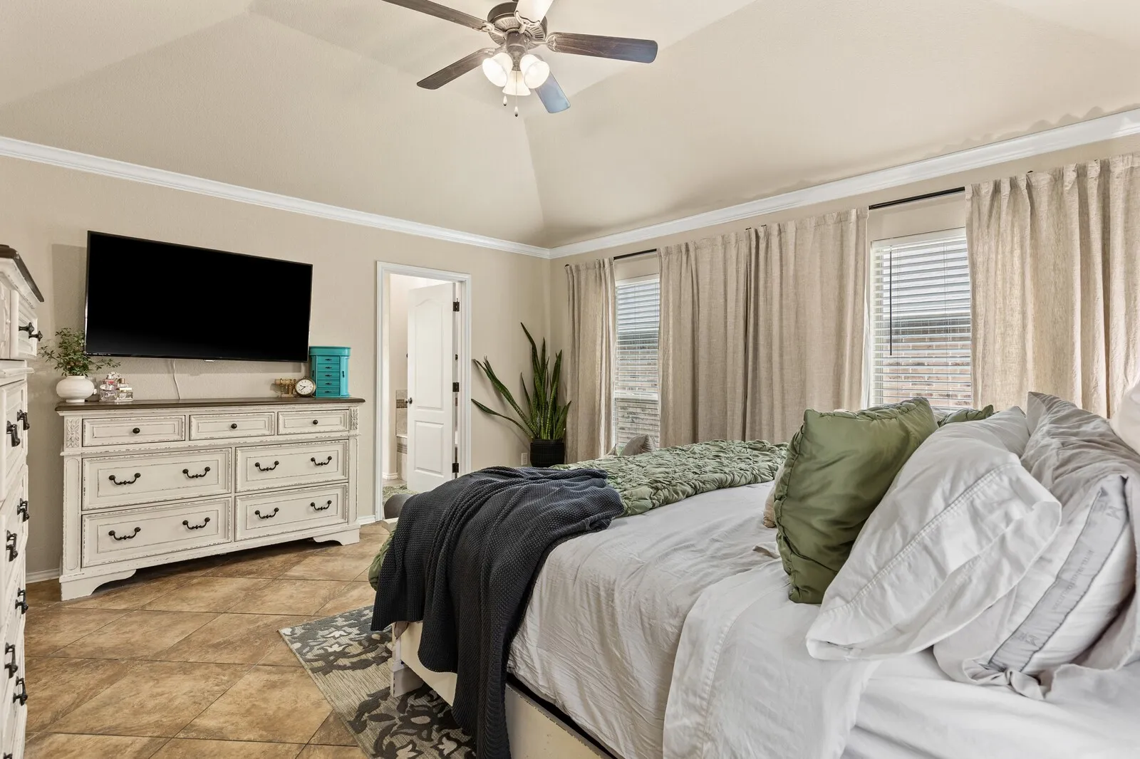 Vaulted ceiling with a ceiling fan and light fixture, featuring crown molding, multiple windows with blinds and drapes, and tile flooring