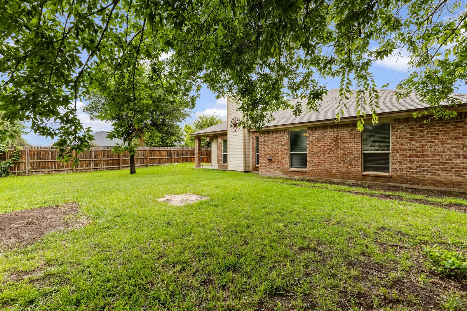 Spacious backyard featuring a full privacy fence, mature trees providing natural shade, a brick exterior, and multiple windows