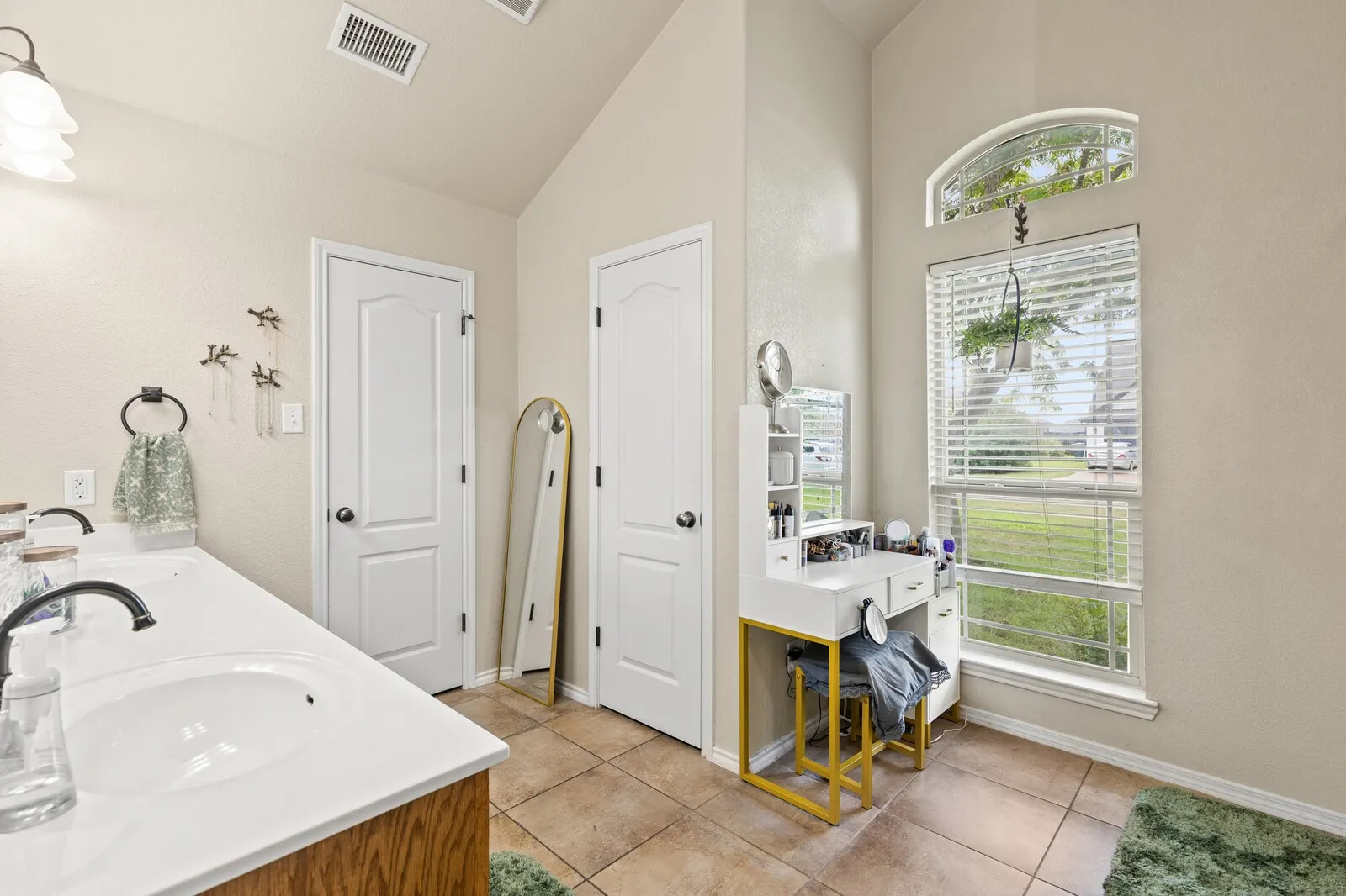 Vaulted ceiling bathroom featuring a dual vanity, light-toned tile flooring, and a large window with an arched transom