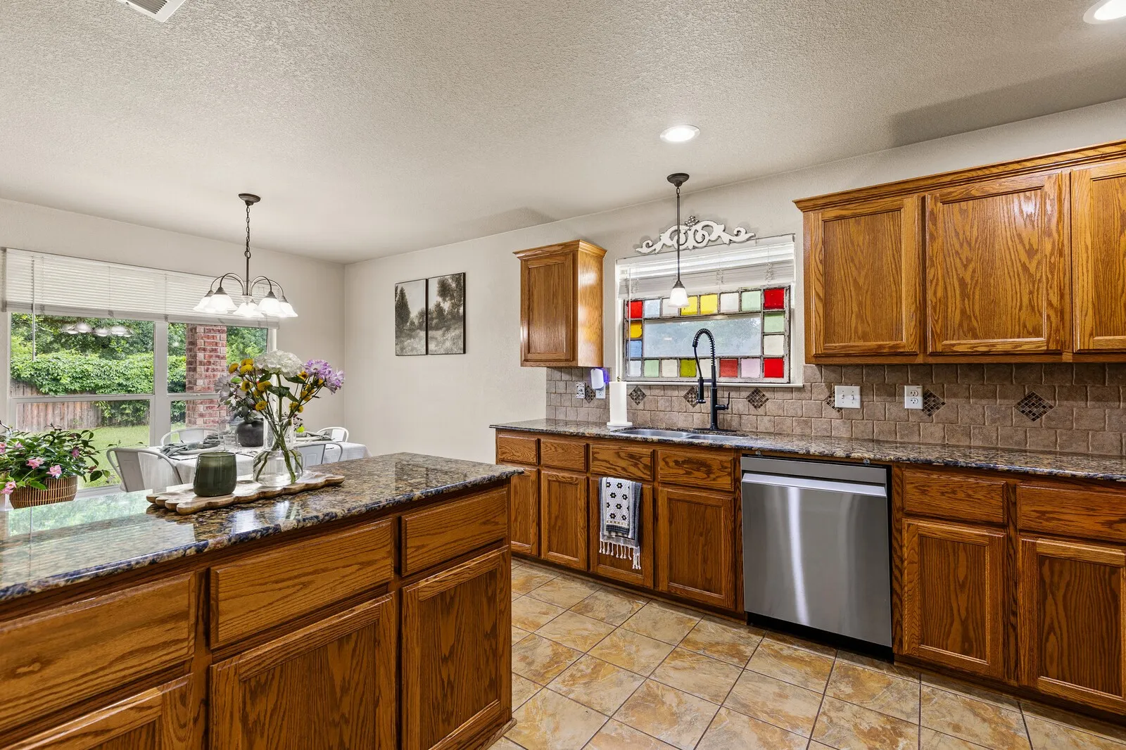 Kitchen featuring granite countertops, wood cabinetry, tile flooring, a stainless steel dishwasher, and a decorative stained-glass window panel above the sink