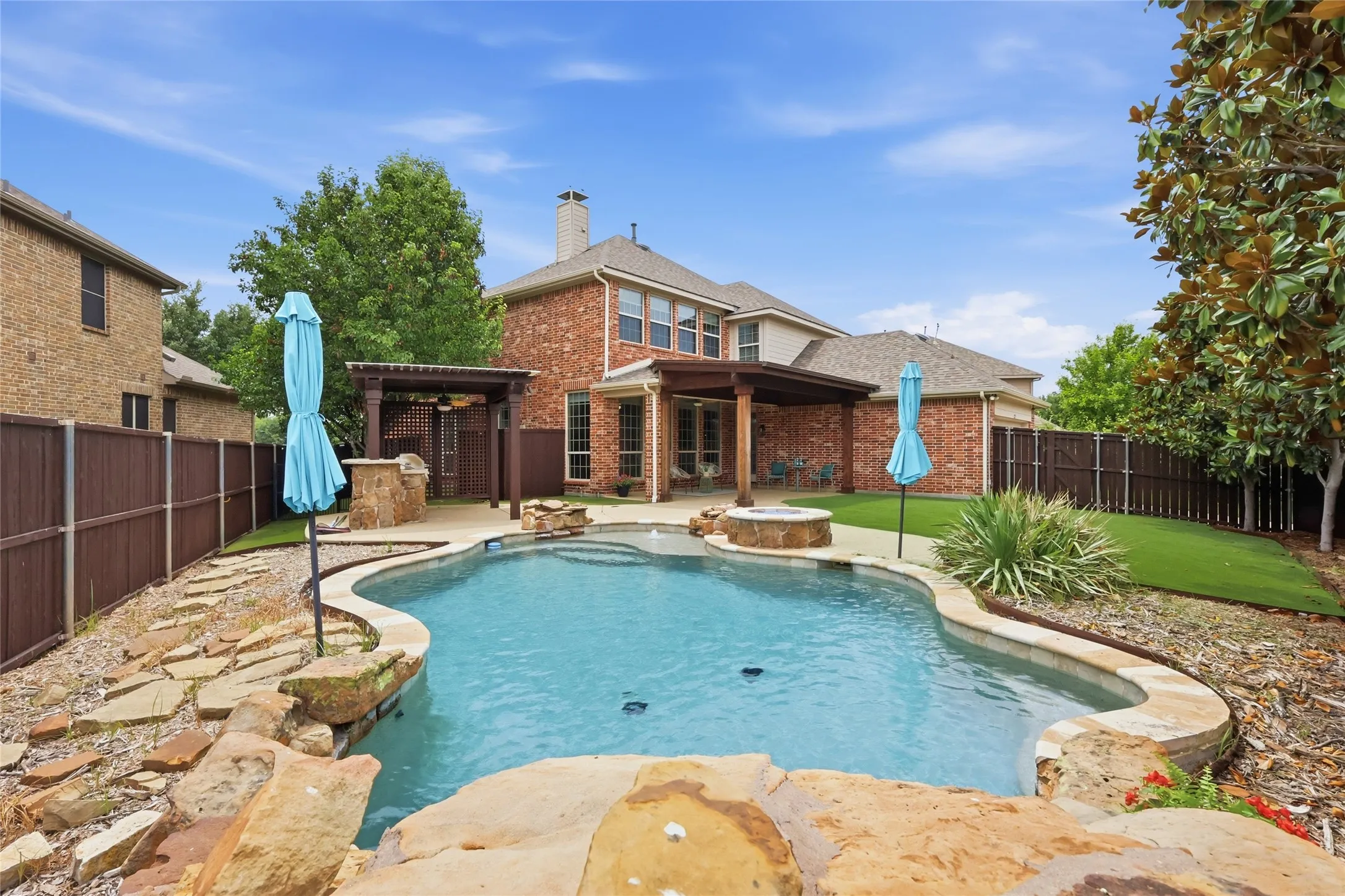 Curved swimming pool with stone coping, a built-in fire pit, covered patio with wood pergola, and an outdoor kitchen area