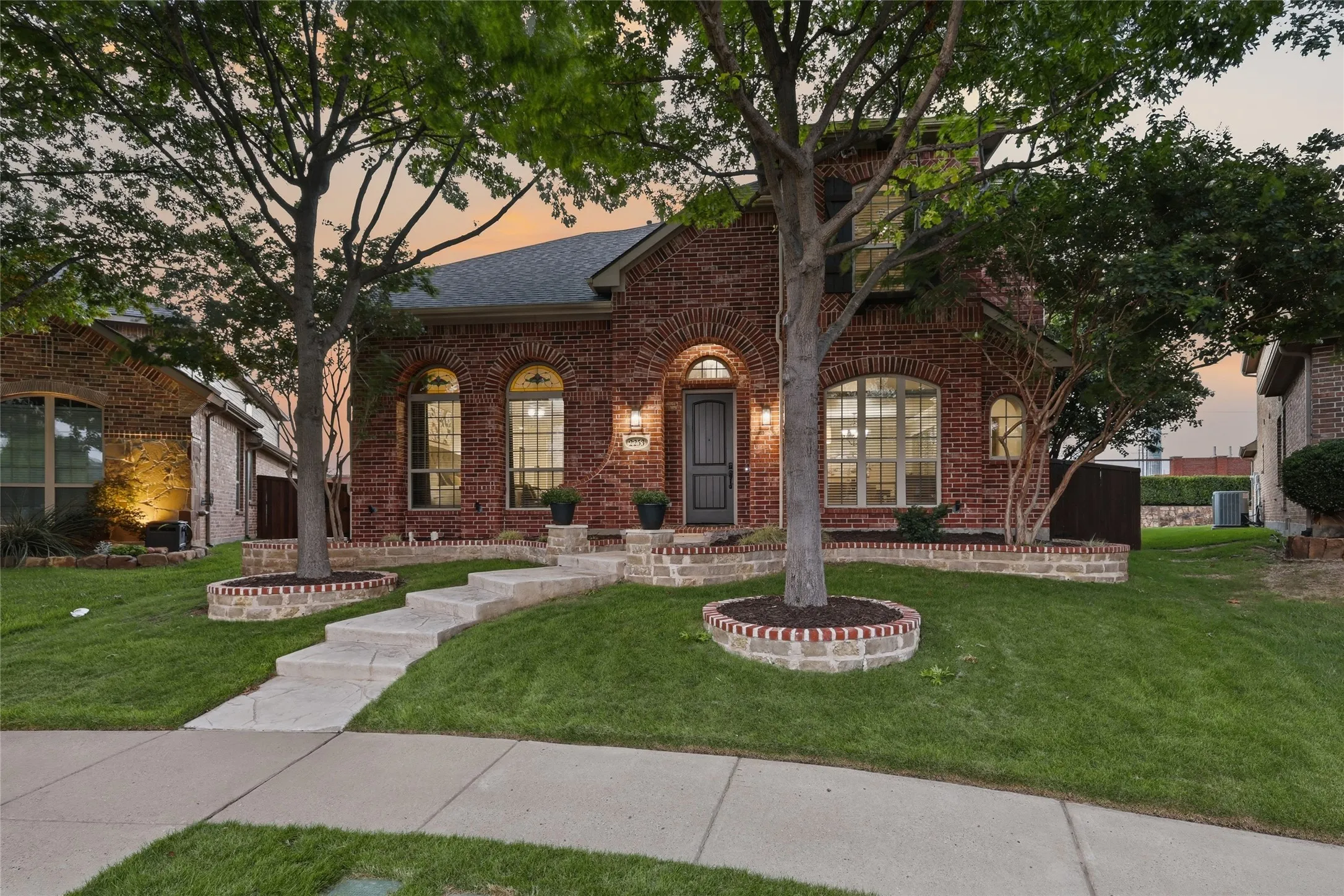 Brick exterior with arched window details, stone-edged landscaping, mature trees, and a concrete pathway leading to the entry