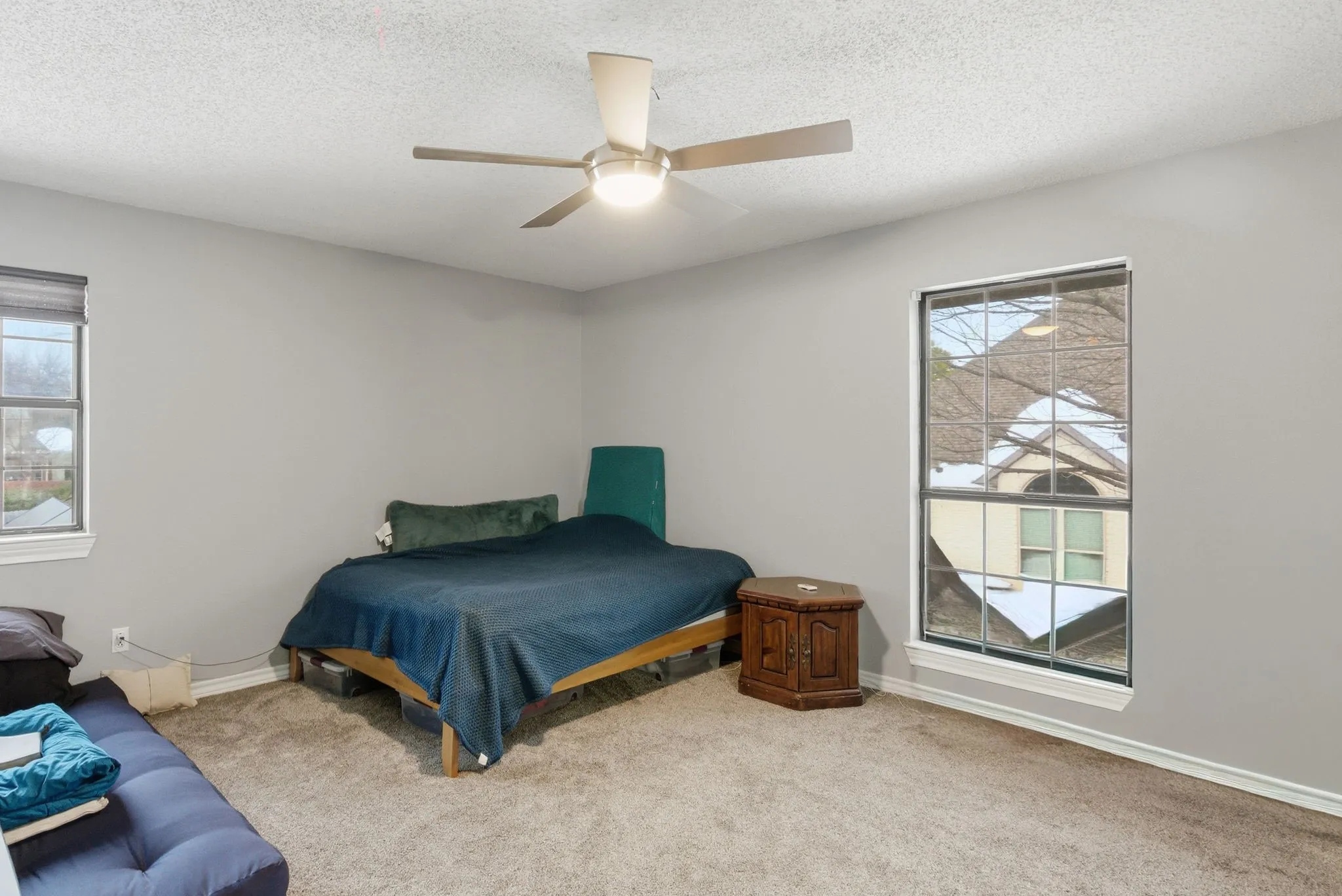 Spacious bedroom featuring light gray walls, textured carpeting, a contemporary ceiling fan with integrated lighting, and two windows with white trim
