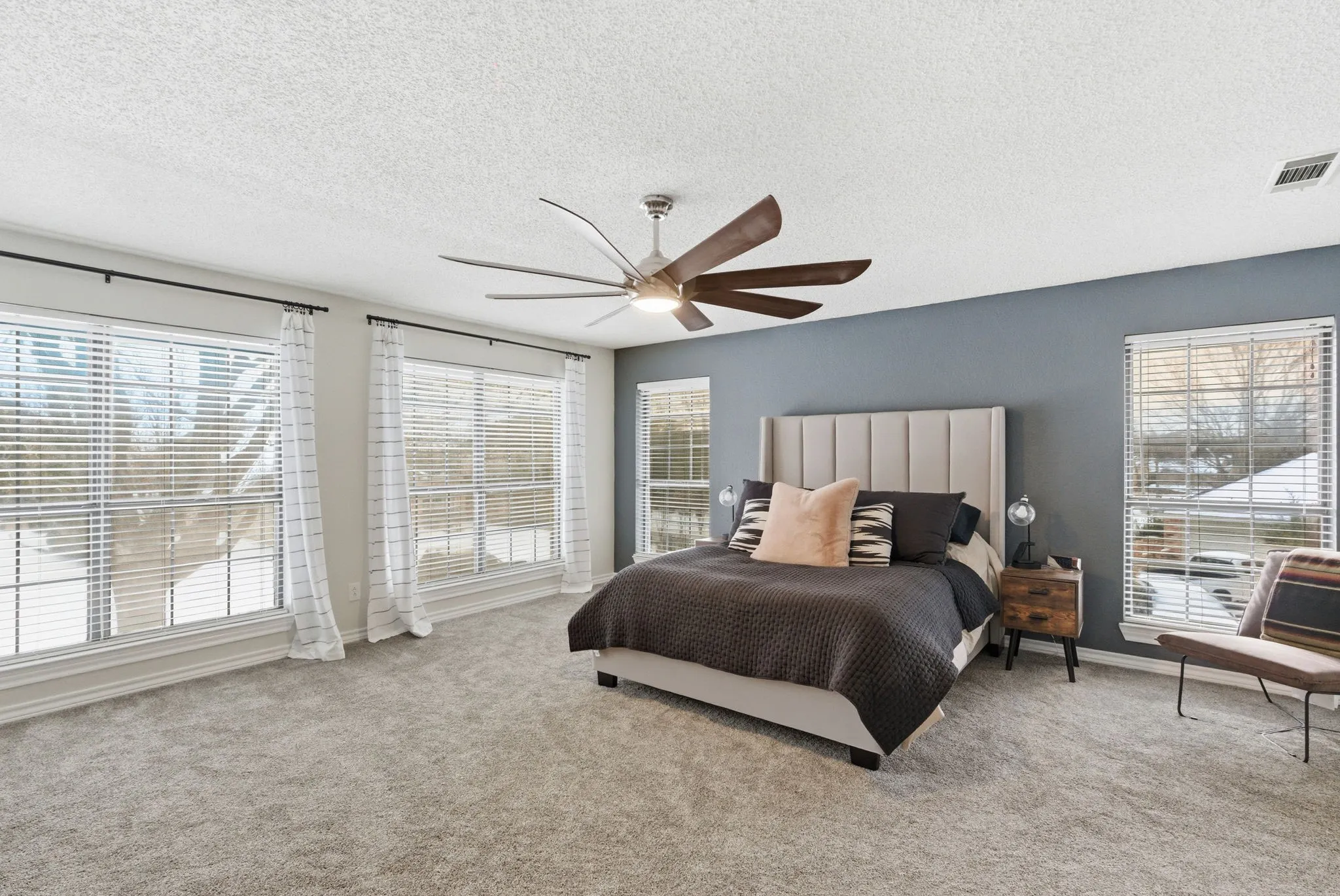 Spacious room featuring light gray carpeting, a multi-blade ceiling fan, and multiple windows with white blinds