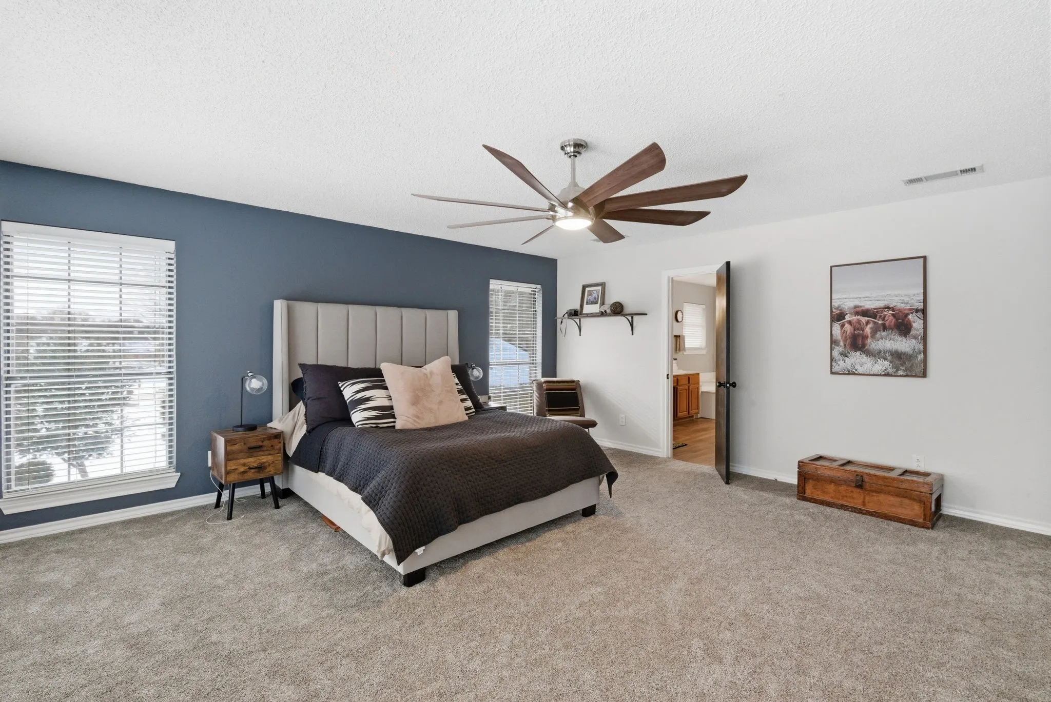 Spacious room featuring carpeting, a deep blue accent wall, two windows with blinds, a ceiling fan with light, and an open doorway revealing a partial bathroom beyond