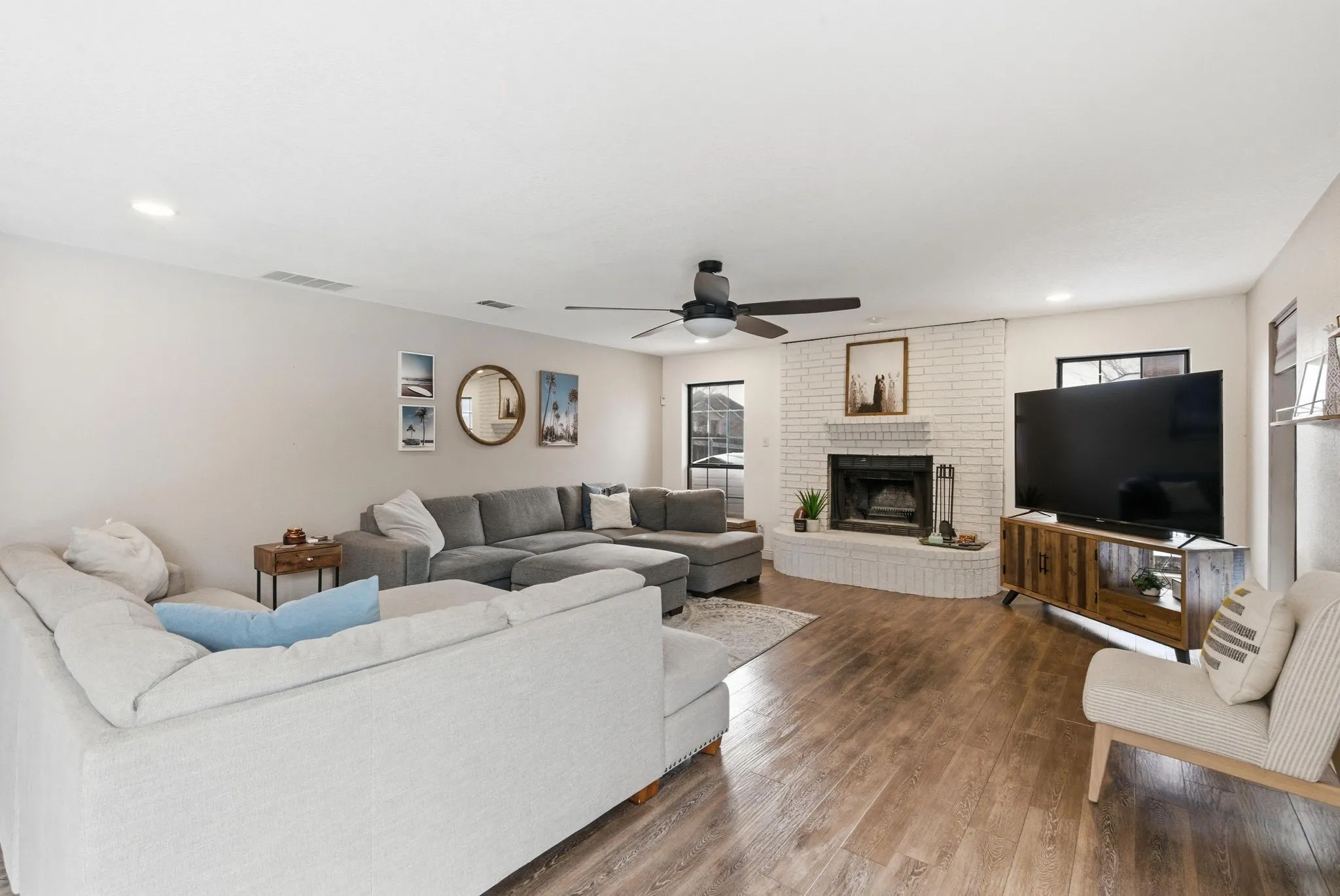 Spacious living area featuring wood-finish flooring, a white-painted brick fireplace, a ceiling fan with integrated lighting, recessed lighting, and wall-mounted windows