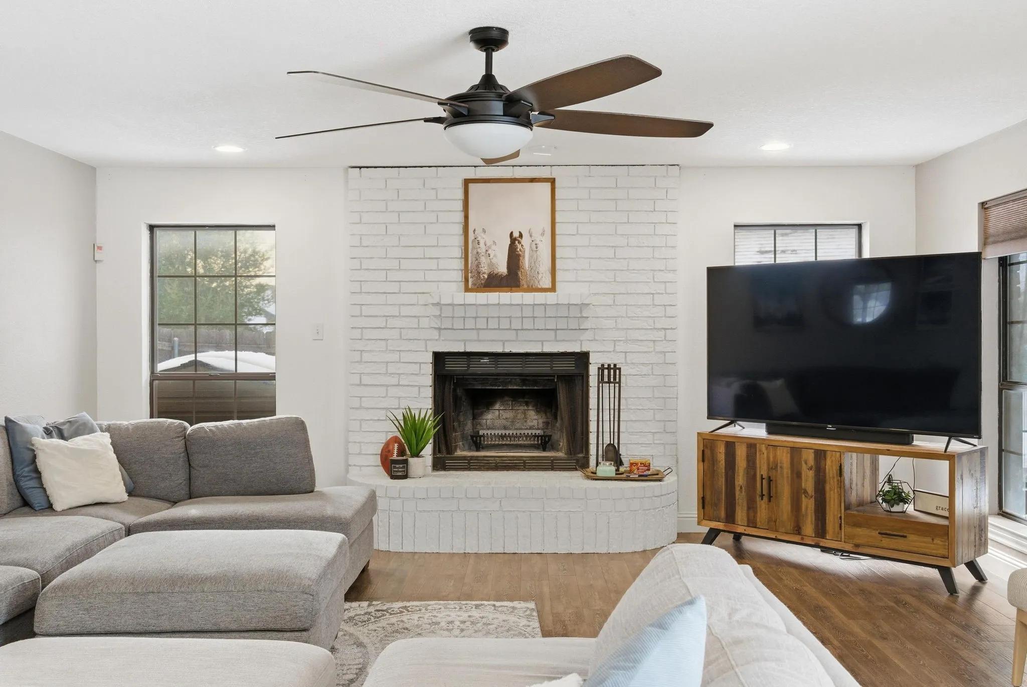 Spacious living area featuring wood-finish flooring, a white-painted brick fireplace, and a ceiling fan