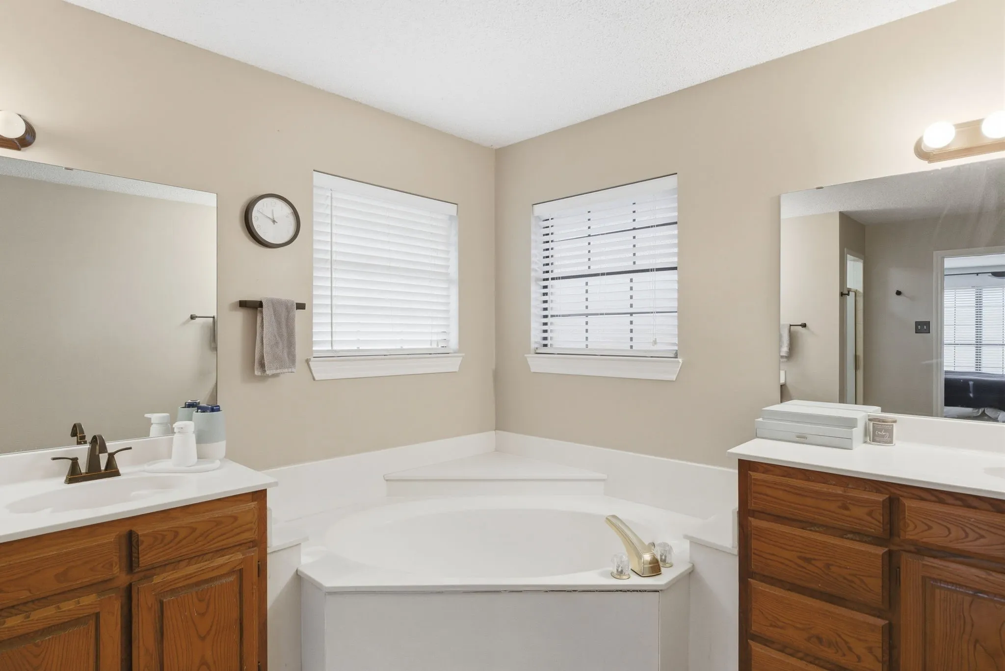 Corner built-in tub with adjacent windows, dual wood-finish vanities with integrated sinks, and individual wall-mounted mirrors