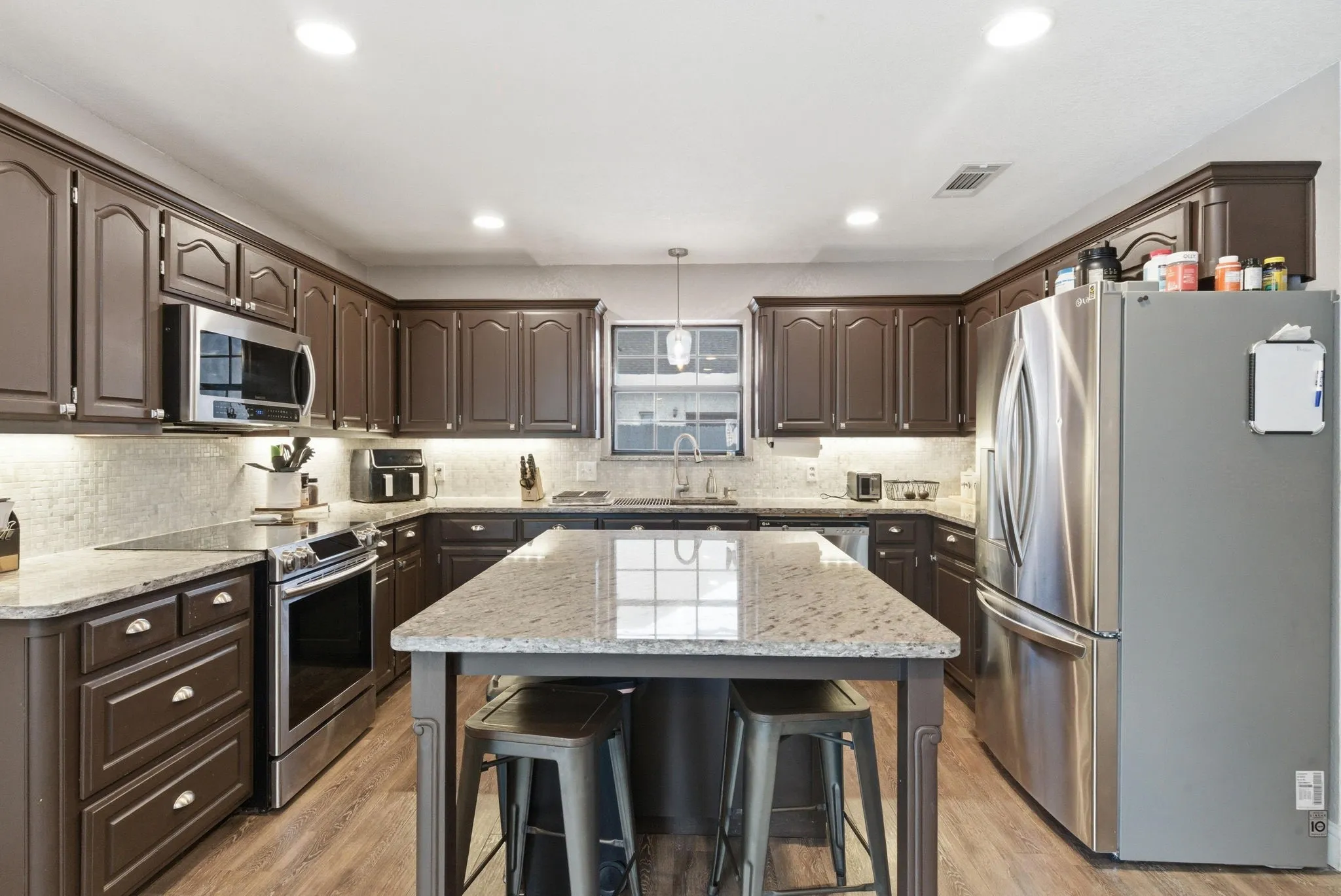 Kitchen featuring an expansive island with a light-toned countertop, extensive cabinetry, stainless steel appliances, a tiled backsplash, and wood-finish flooring