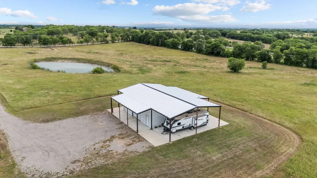 Expansive property featuring a large metal building with an attached covered concrete pad, a gravel driveway, and a pond surrounded by green fields