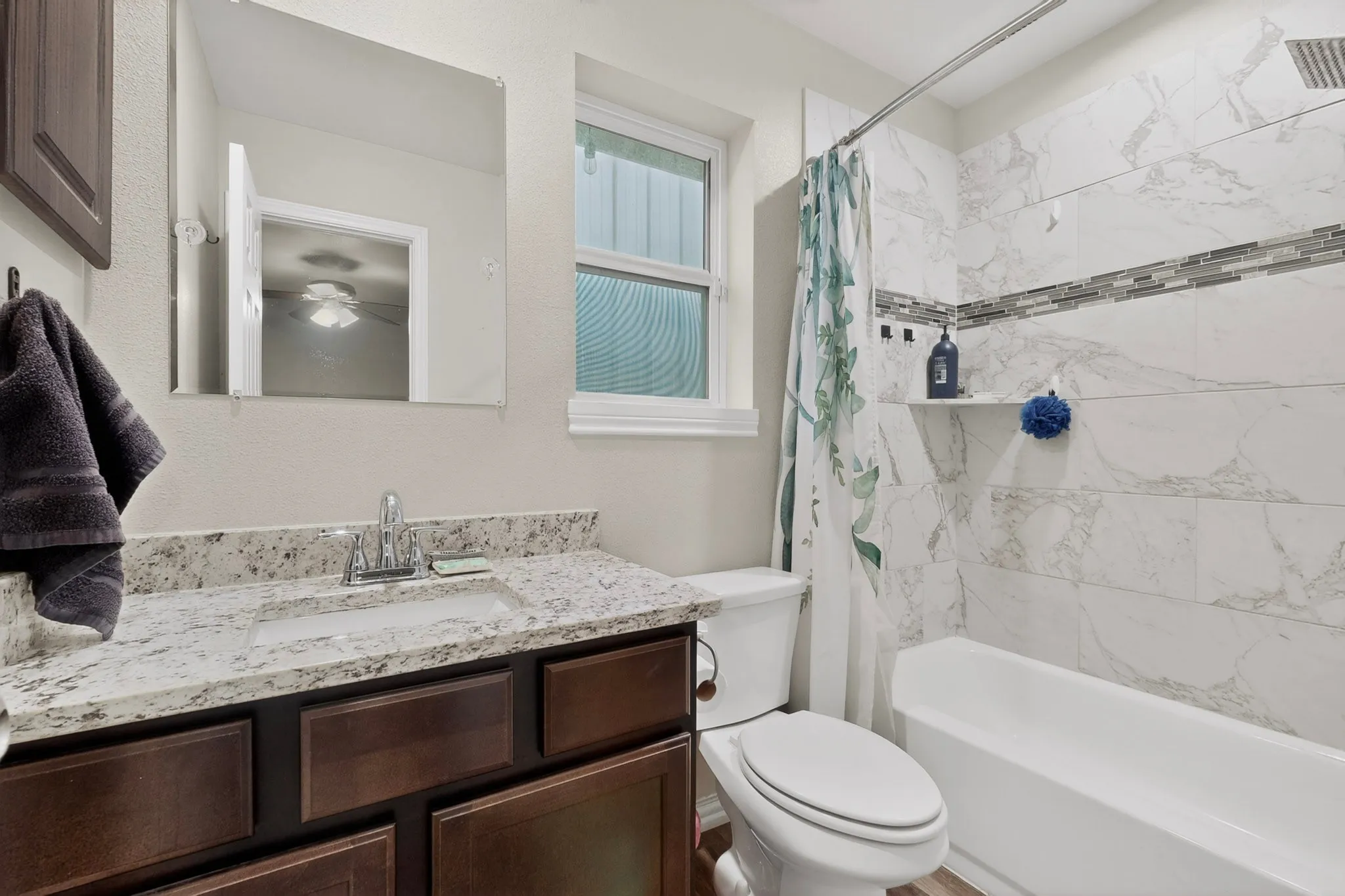 Bathroom featuring a dark wood vanity with a light-colored speckled countertop, an undermount sink with a chrome faucet, a white toilet, and a bathtub with a marble-look tiled surround and a mosaic accent band