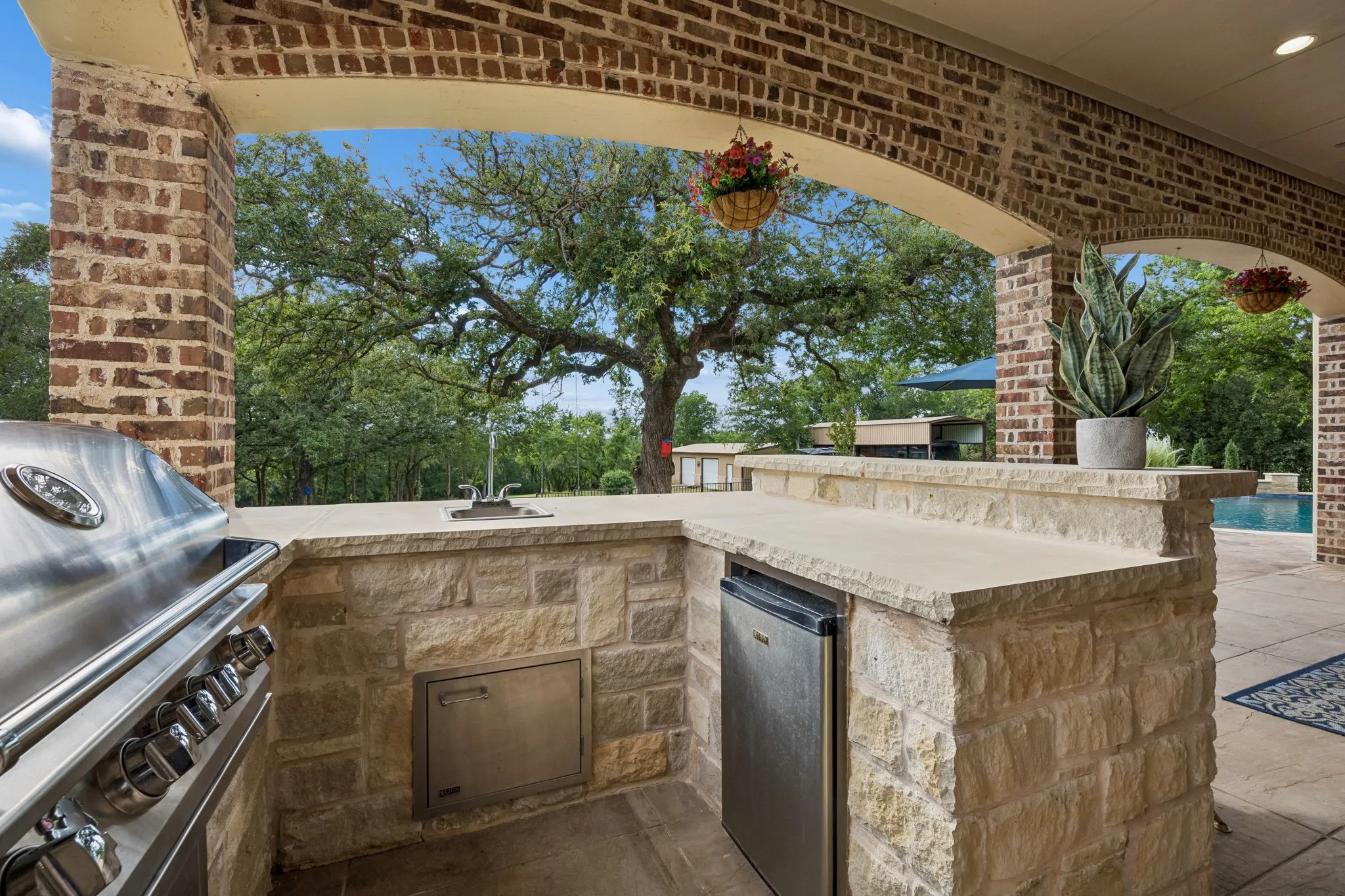 Outdoor kitchen featuring a built-in stainless steel grill, integrated sink, and under-counter refrigerator within a stone-clad structure