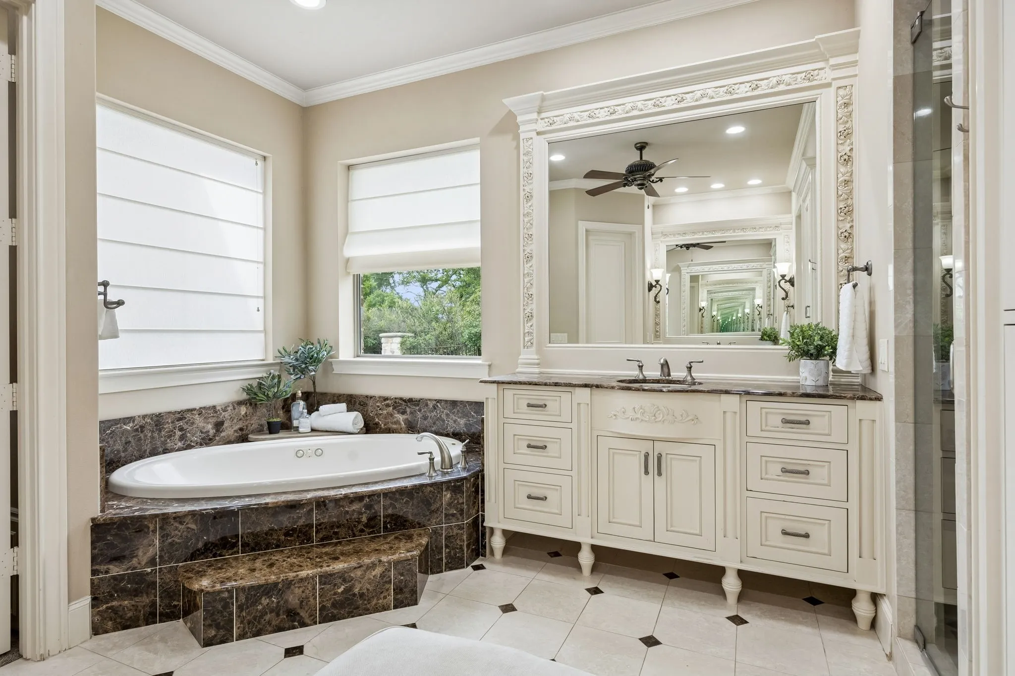Soaking tub with dark marbled tile surround, dual vanity with decorative carved mirror, light cabinetry with bronze hardware, glass-enclosed shower, and checkered tile flooring