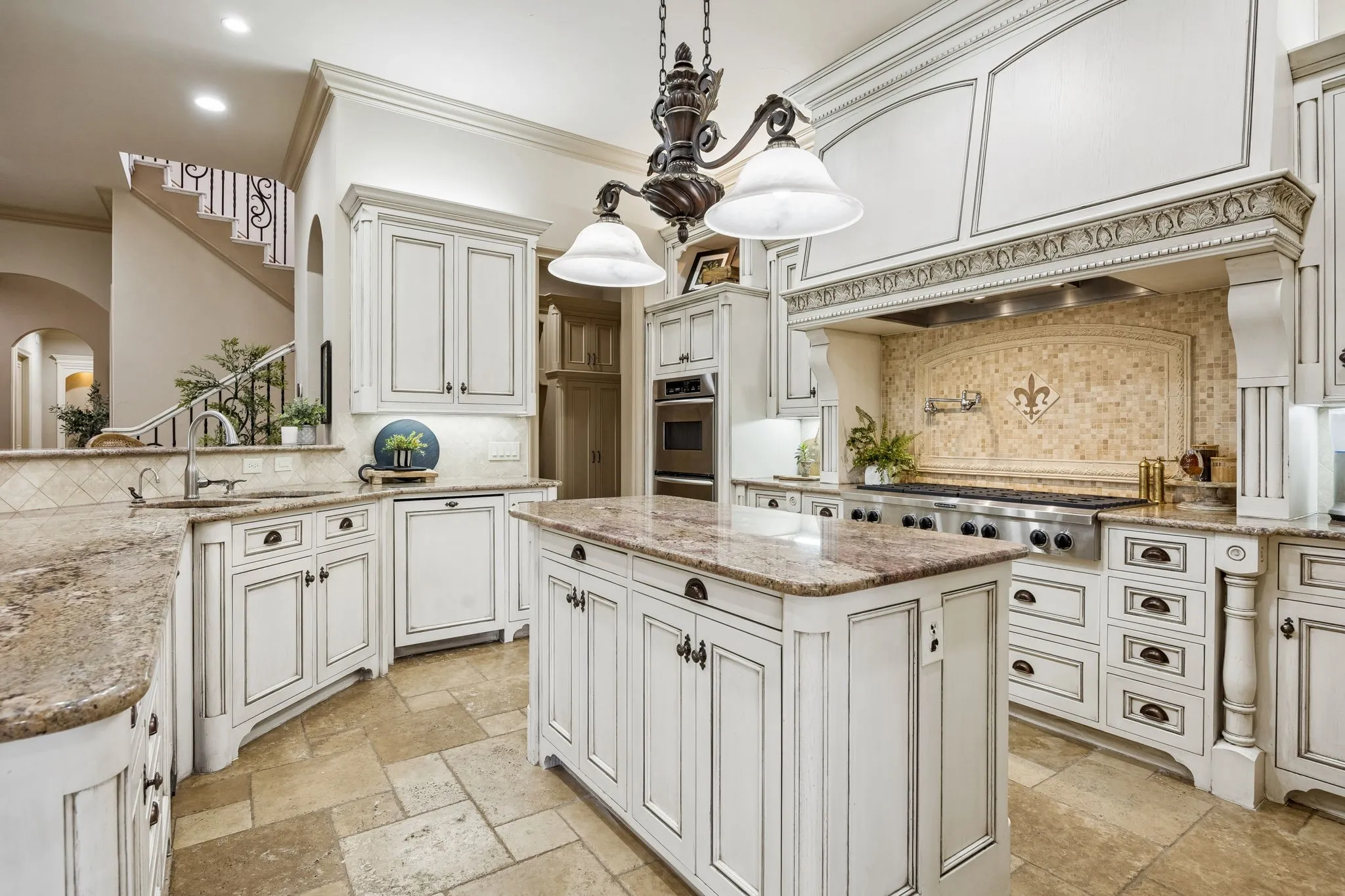 Gourmet kitchen featuring a large central island, stone tile flooring, custom cabinetry with ornate details, built-in stainless steel appliances, and a decorative range hood with a tiled backsplash