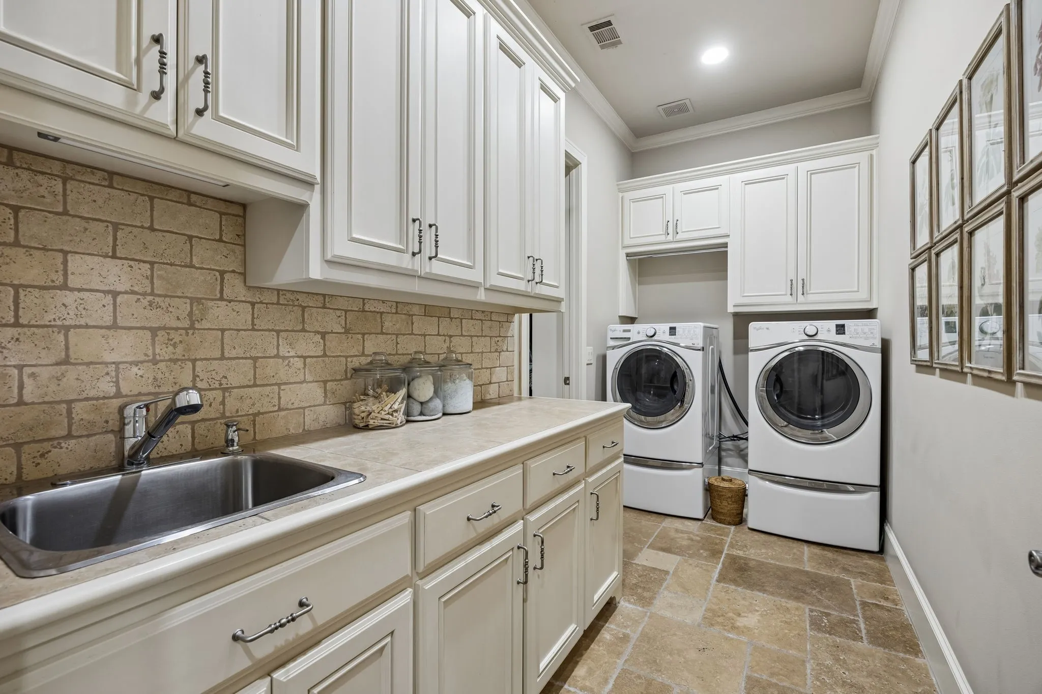 Dedicated laundry space featuring extensive white cabinetry, a utility sink, a stone-look countertop, brick-patterned tile backsplash, and large format floor tiling