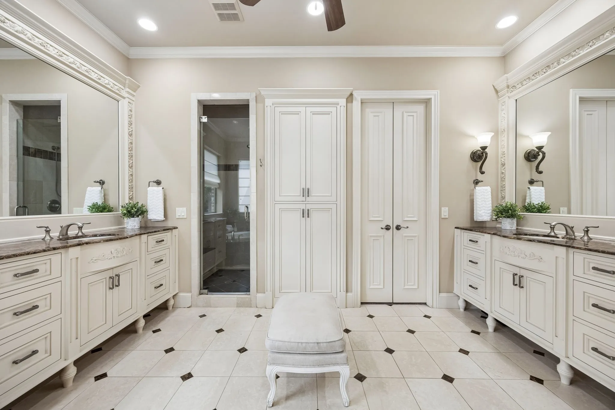 Spacious bathroom featuring two separate vanity areas with stone countertops and decorative mirrors, a glass-enclosed shower, built-in storage cabinetry, and a patterned tile floor