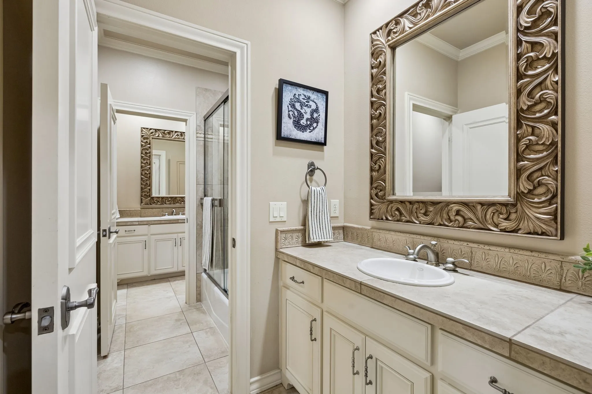 Full bathroom featuring a single vanity with an oval undermount sink, a large ornate framed mirror, a tiled countertop, and white cabinetry with dark hardware